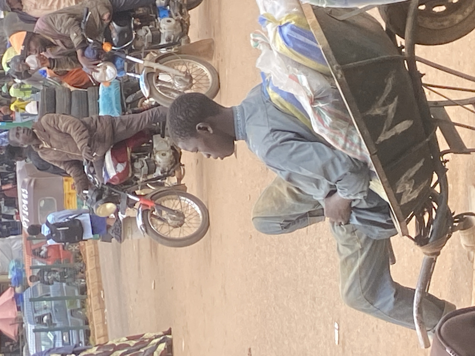 The image depicts a busy street scene in Bukuru, Nigeria. The primary subjects are several individuals, some of whom appear to be vendors or people transporting goods. In the foreground, a young person wearing a light blue shirt and pants is pushing a cart laden with various items, possibly for sale or transport. The cart has a dark metal frame and is filled with bundled goods, some wrapped in plastic and fabric with patterns of yellow, blue, and white.
Behind this individual, a person in a brown jacket is seated on a motorcycle. This motorcycle, along with another one visible further back, suggests a mode of transportation and possibly a marketplace environment. The motorcycles are common in this region for both personal use and as a means of carrying goods or passengers.
The setting appears to be an outdoor marketplace or a busy street. The ground is a sandy, earthy color, consistent with the terrain in many parts of Nigeria. The lighting suggests it is daytime, likely morning or afternoon, with the sun casting some shadows. The overall atmosphere seems active and bustling, typical of a local market.
Notable details include the license plate on a vehicle in the background, which reads "PT10445". Various other vehicles, including a tricycle and what appear to be minibuses, are also present, indicating a developed transportation hub. Several colorful fabrics and umbrellas are visible, adding to the vibrant market feel. The people in the image are dressed in everyday clothing suitable for the warm climate. There are no strong emotional cues visible, but the activity suggests a sense of purpose and daily life.