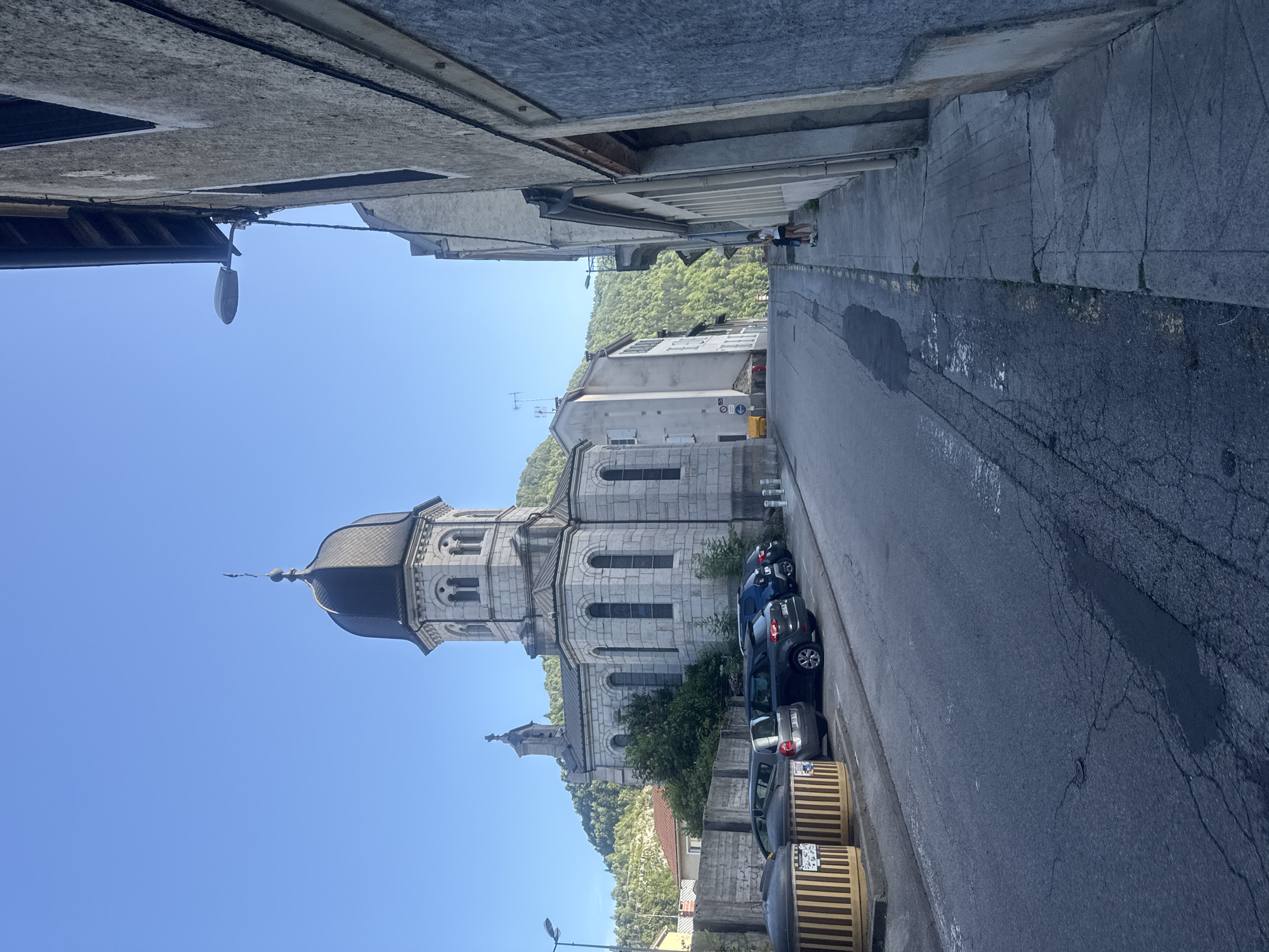 The image depicts a street scene in the city of Saint-Claude, France, dominated by a stone church with a dark, domed roof and a tall steeple. The church is situated on a slight incline, with a row of parked cars on the left side of the street in front of a concrete barrier and some large cylindrical trash receptacles. The street itself is paved and shows signs of wear, with cracks and some wet patches.
To the right of the street, a sidewalk runs alongside a textured, light-colored building. A street lamp is mounted on the side of this building. Further down the sidewalk, two individuals, a child and an adult, are visible. The adult appears to be wearing a white top and shorts, and the child is wearing dark shorts and a blue shirt. They are standing and facing away from the camera.
Beyond the immediate street view, more buildings are visible, including some with red roofs, and a densely forested hillside rises in the background. The sky is clear and blue. The overall impression is a quiet, daytime street in a town with architectural elements suggesting historical significance.