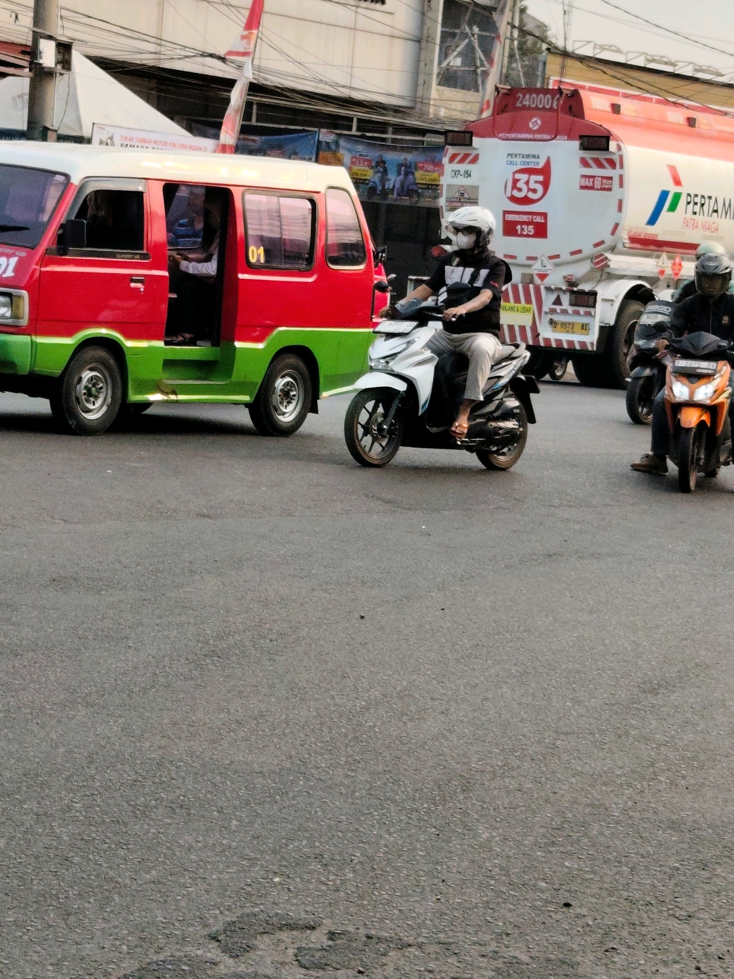 In Purwakarta, Indonesia, a traffic scene unfolds. A red and green van, possibly a public transport vehicle, is stopped with its doors open. A person sits in the driver's seat, while another appears to be standing near the open passenger door. In the background, a large Pertamina tanker truck is visible, displaying "24000 L" and "35 EMERGENCY CALL 135." Two motorcyclists are in the foreground. One, wearing a white helmet and face mask, rides a white scooter. Another motorcyclist on an orange scooter is partially visible to the right. The road is paved and appears to be an urban street, with overhead wires and some indistinct buildings in the distance. The lighting suggests it is daytime.