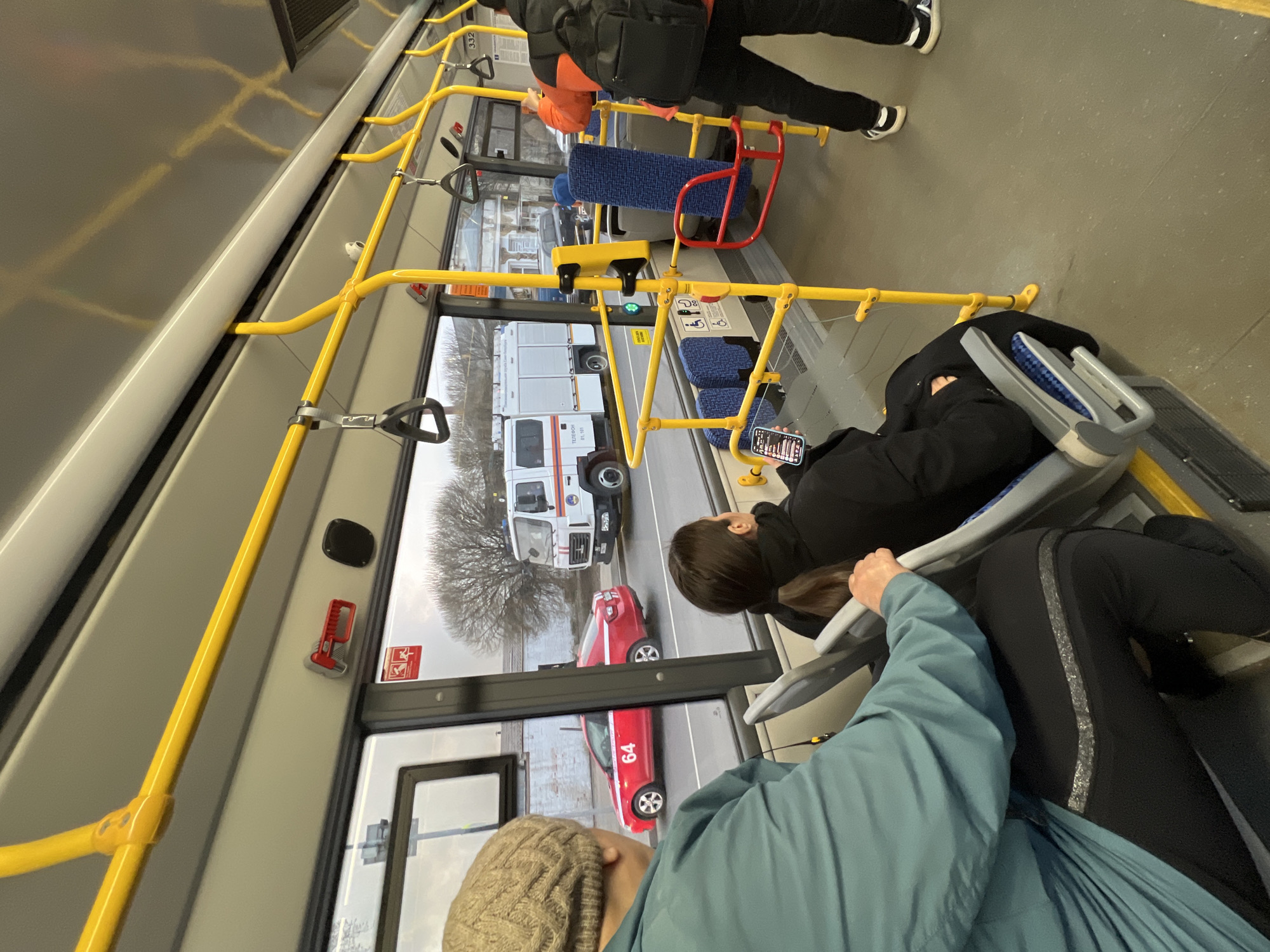 The image depicts the interior of a bus, likely in Saint Petersburg, Russia. Two red cars, one of which has a number 64 on it, and a white vehicle are visible through the bus windows. The interior of the bus has yellow handrails. Several passengers are on board, and some are seated.