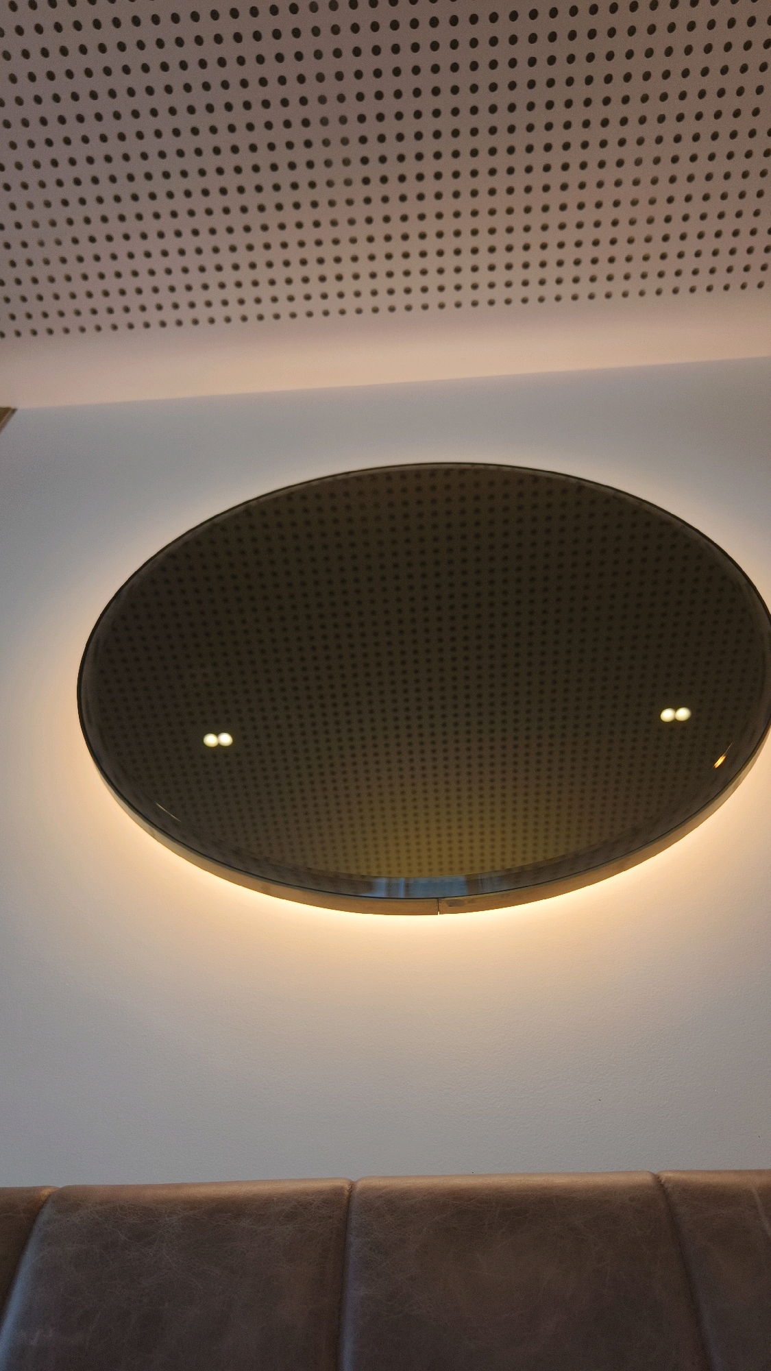 An oval-shaped, dark, perforated surface is illuminated from behind by a warm, orange light. This surface is mounted on a white wall, above a brown, upholstered bench. The ceiling above the oval surface is also perforated, with a grid of small, dark dots. The image appears to be a detail shot of interior design, possibly in a restaurant, cafe, or lounge. The specific location in Vienna, Austria, cannot be determined from the image alone.