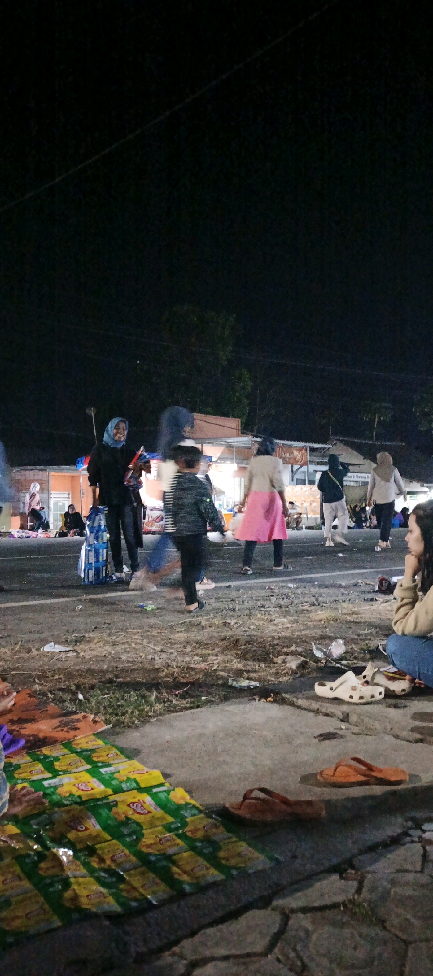 The image depicts a nighttime scene in Kasiyan, Indonesia, likely at a roadside market or gathering. In the foreground, a mat of snack packaging is laid out on the ground, accompanied by several pairs of flip-flops and a white clog. Further back, people are visible walking and standing. Some individuals are wearing traditional Islamic head coverings. A stack of blue and white bags is also present. The background shows structures that appear to be stalls or small shops, illuminated by artificial light, with trees and a dark sky above. The ground is a mix of asphalt and rougher terrain, with scattered debris. The overall impression is of a lively, albeit somewhat disorganized, public space at night.