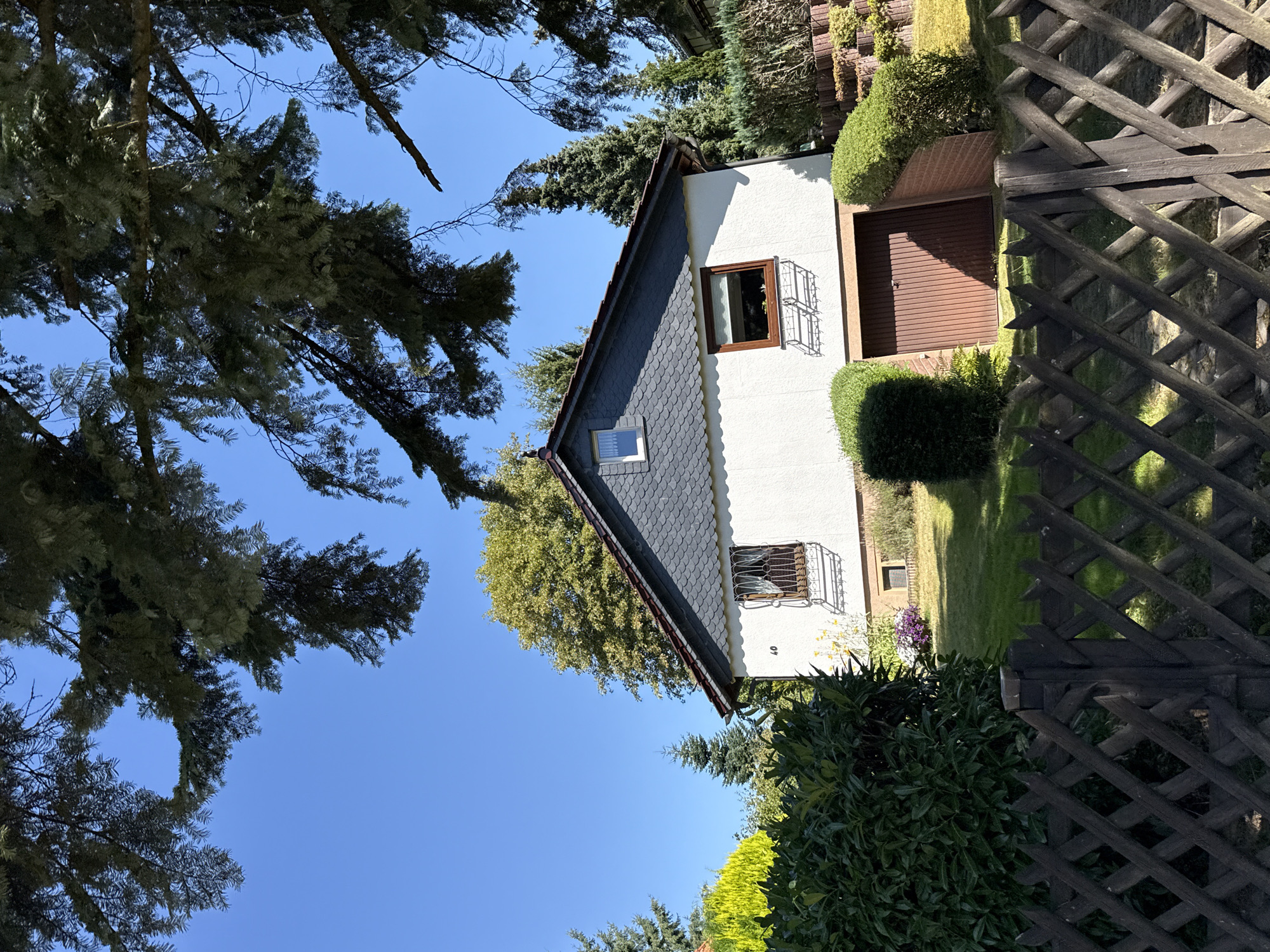 A white stucco house with a dark gray, scaled shingle roof is situated in a suburban setting in Bad Sachsa, Germany, under a clear blue sky. The house features two windows on its visible facade, one with a decorative wrought-iron grill and the other a smaller square window in the gable. A brown wooden garage door is visible to the right of the main house entrance. The property is bordered by a dark wooden, diamond-patterned fence in the foreground. Lush green bushes and trees surround the house, with a mature tree's branches extending into the upper frame of the image, casting shadows. The lawn is well-maintained.