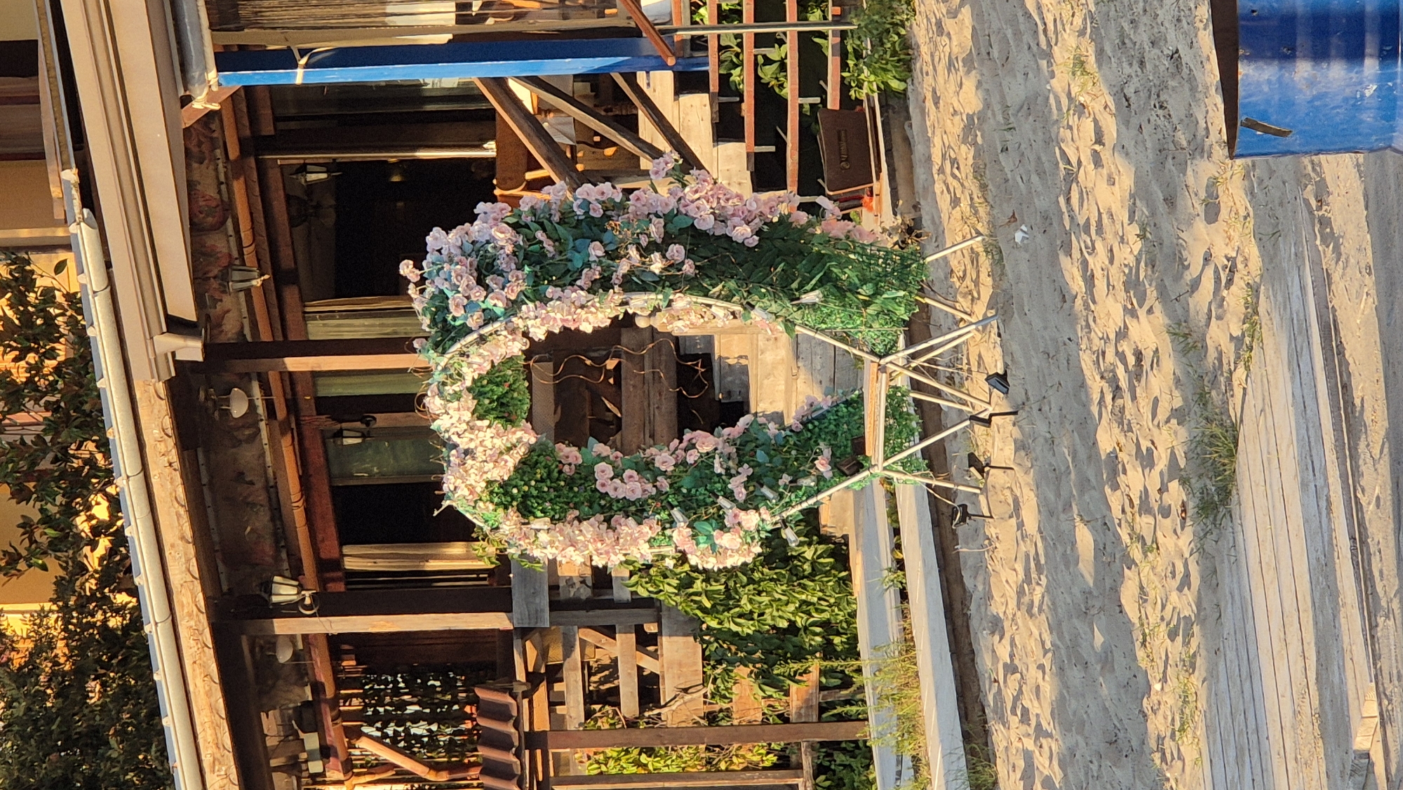 The image depicts an outdoor setting, likely a beach or a waterfront area in Kiten, Bulgaria, characterized by sandy ground and wooden structures. Dominating the center is a large, heart-shaped archway, elaborately decorated with artificial green foliage and numerous pink and white artificial flowers. The archway is supported by a white metal frame, partially obscured by the foliage. Two folding chairs are placed inside the base of the arch. Behind the archway, a wooden structure resembling a building with a porch and stairs is visible. The building has wooden pillars and a roof, and its facade features windows and doors. Green plants and foliage are abundant around the wooden structure and on the sandy ground. In the foreground, the sandy beach stretches out, with a section of wooden planks or a walkway. To the far right of the image, a large blue barrel is partially visible. The lighting suggests it is either early morning or late afternoon, with long shadows cast across the sand.