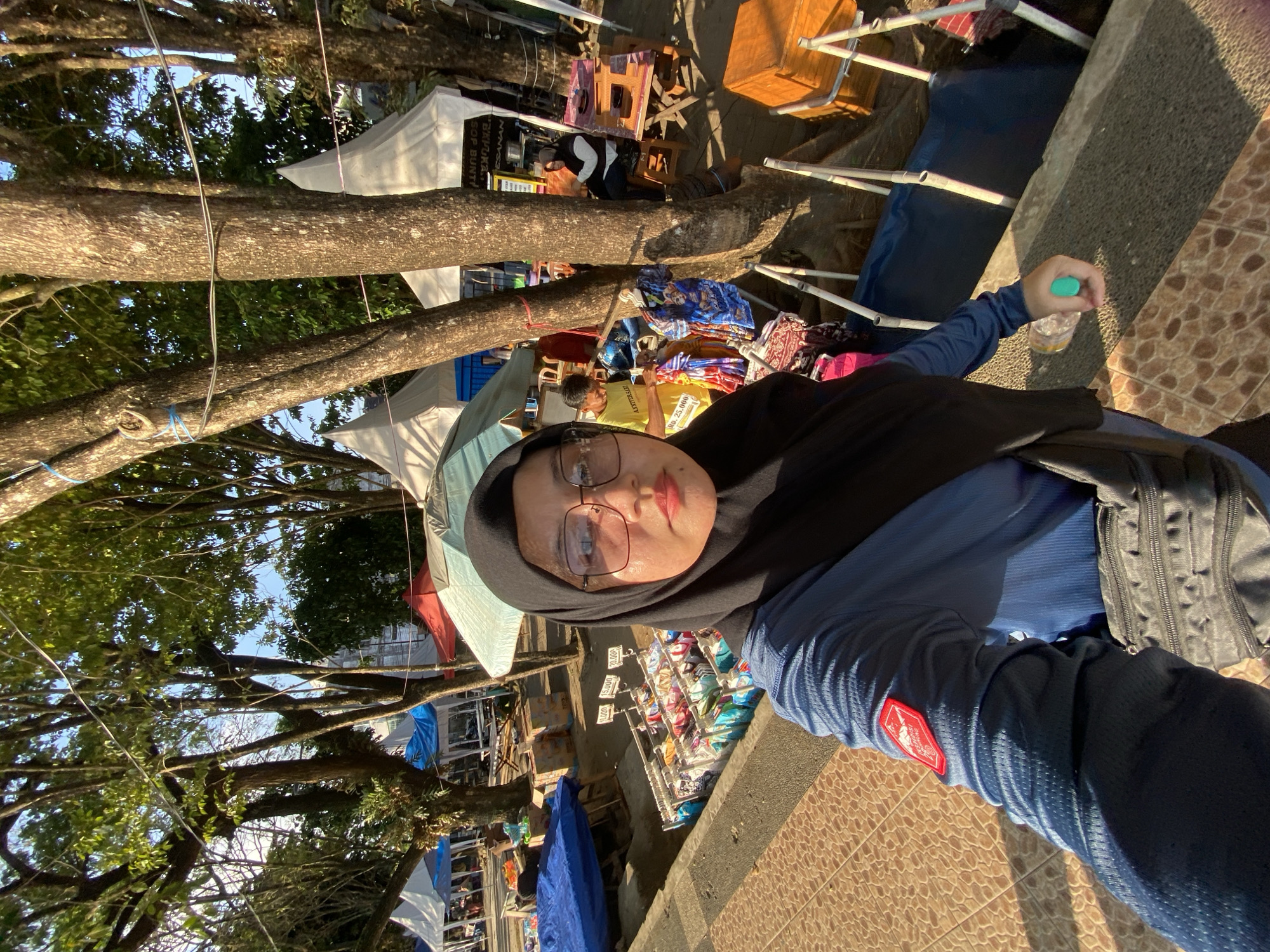 A person in a blue long-sleeved shirt and black hijab, wearing large glasses, takes a selfie outdoors. They are standing on a tiled walkway next to a market stall displaying clothing on hangers. Behind them, tents and trees are visible. The scene appears to be at the Siring Tendean market in Banjarmasin, Indonesia, known for its floating markets and vendors. The time of day is indicated by the sunlight casting shadows.