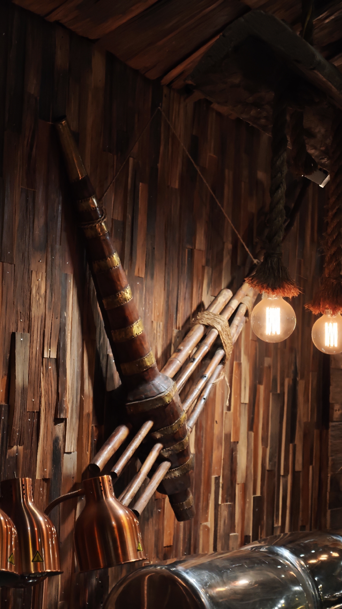 The image shows a restaurant interior in Sa Pa Ward, Vietnam, with a rustic, ethnic-inspired décor. A traditional Hmong musical instrument, likely a Khene, is mounted on a wooden wall. The wall is paneled with narrow strips of wood, arranged vertically. The instrument is made of bamboo pipes bound together and fitted with a wooden or gourd resonator, and decorated with metal bands. To the right of the instrument, two vintage-style light bulbs hang from rope suspensions. In the foreground, the tops of copper-colored food warmers are visible, reflecting light. The lighting is dim, with warm tones.
