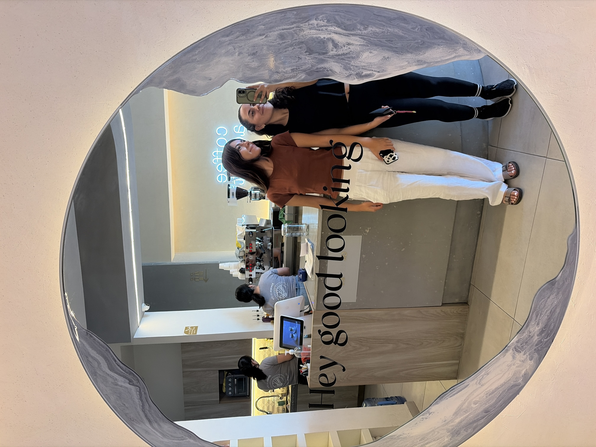 This image, rotated 90 degrees clockwise, features a mirror selfie taken by two women inside what appears to be a modern cafe or coffee shop. The central element is a large, irregularly shaped mirror with a distinctive wavy, grey-marbled border that frames the reflection.
Two women are posing in front of the mirror. The woman on the right is dressed in a black short-sleeved top, black leggings, and black athletic shoes with white-striped socks. She holds a dark-colored smartphone up to capture the selfie, looking towards the camera with a subtle smile. Beside her, the second woman wears a rust-brown short-sleeved top, white wide-leg pants, and open-toed sandals. She holds a smartphone with a black and white cow-print case in her left hand. Both women have dark hair and are looking upwards with relaxed expressions.
The reflection in the mirror reveals the interior of the establishment. A light blue neon sign spelling "coffee" is visible behind the women. Further in the background, a counter area is equipped with coffee-making machinery, including an espresso machine, various cups, bottles, and a tablet-style point-of-sale system. Two staff members, wearing grey t-shirts (one with a visible but indistinguishable logo), are seen working behind the counter. The phrase "Hey good looking" is written in large black letters across the surface of the mirror, partially obscured by the women's figures. The setting suggests a clean, contemporary commercial space.