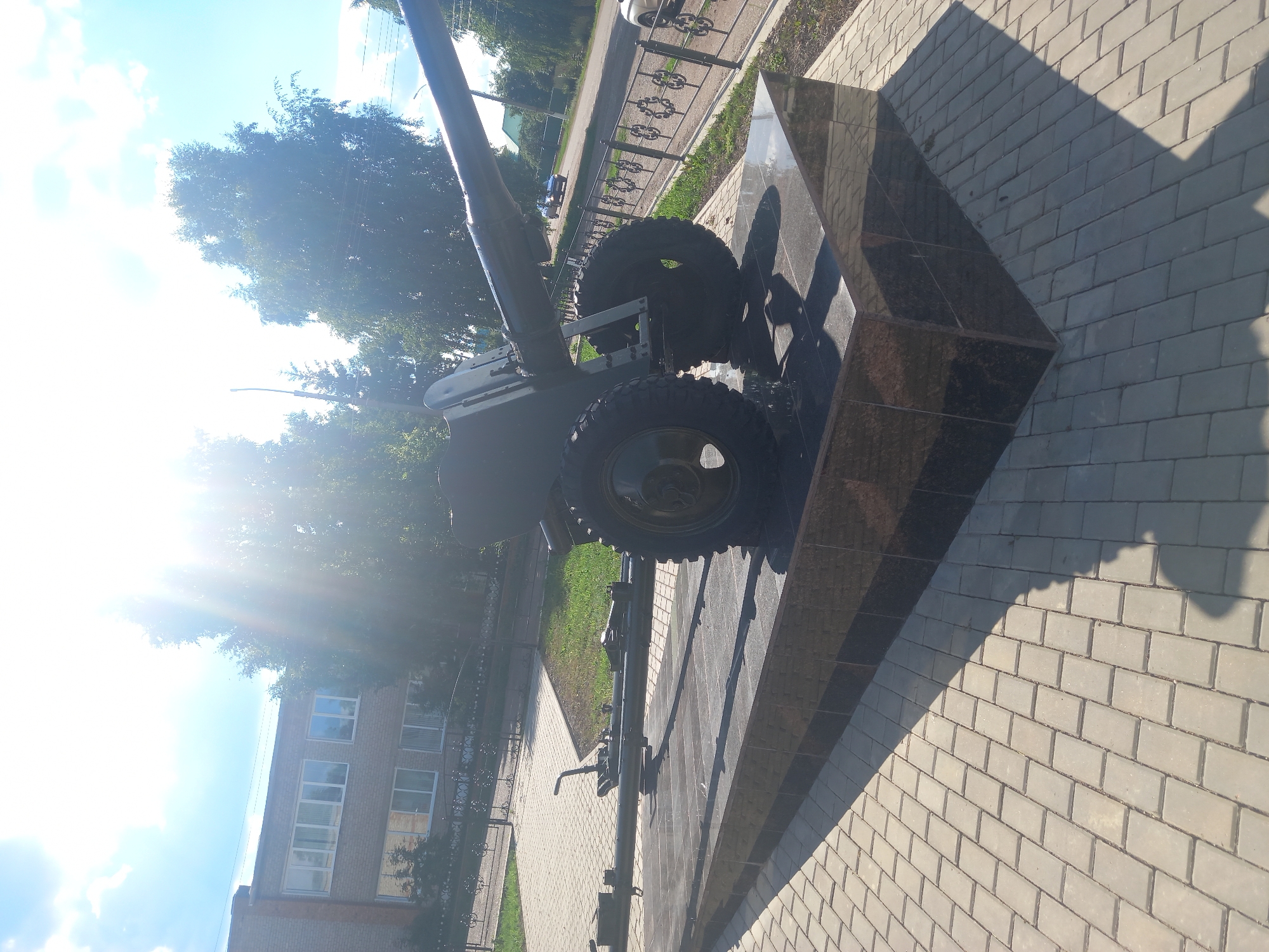 The image depicts an outdoor scene with a monument to a cannon situated on a paved area. The monument rests on a dark, polished stone base. The cannon is dark green and features two large wheels. To the left of the cannon, a portion of another similar structure is visible. The background includes a brick building with multiple windows, trees, and a street with a fence. The sun is bright, casting shadows and creating lens flare effects. The location is identified as Sterlibashevo, Russia.