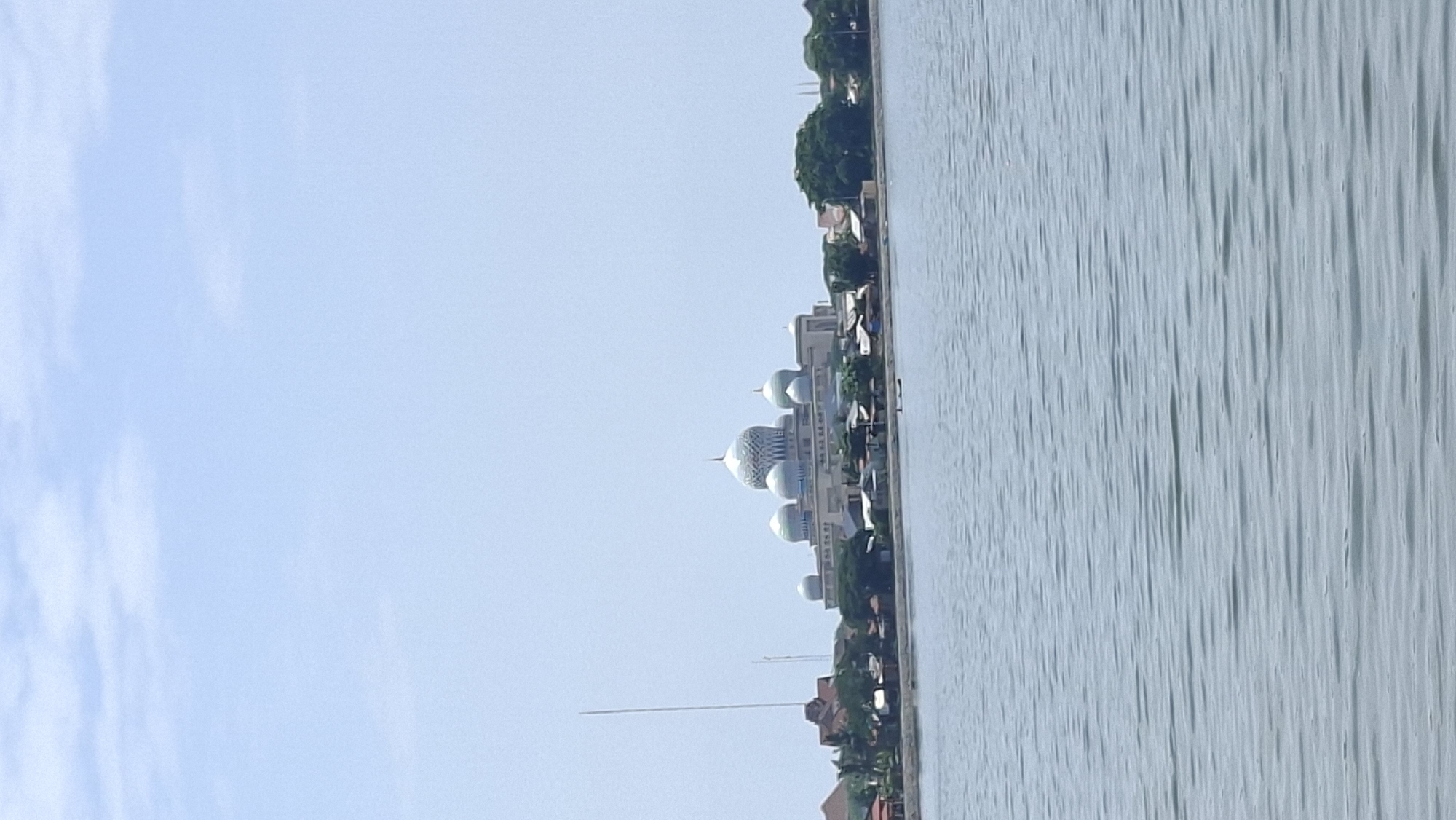 The image shows a wide shot of a body of water in front of a mosque and surrounding buildings, likely in the city of Lhokseumawe, Indonesia. The mosque is the dominant structure, featuring multiple domes and a tall minaret-like tower. The water is calm with visible ripples, reflecting the overcast sky. Several smaller buildings, trees, and a tall antenna structure are visible on the shoreline behind the mosque. A couple of small boats are present on the water. The scene suggests a daytime view under a bright but cloudy sky.