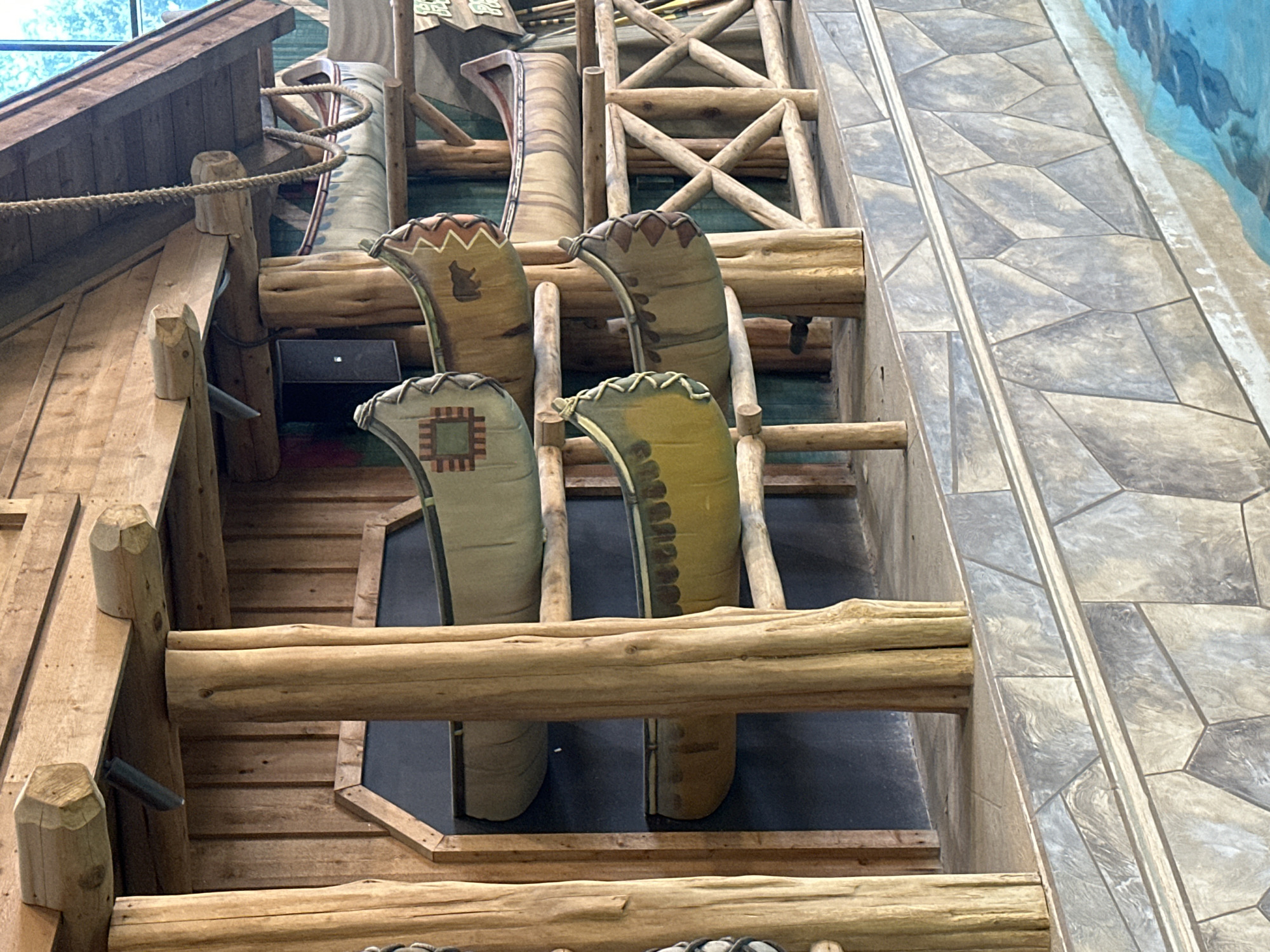 A decorative display of canoes is mounted on a wooden structure, suggesting an indoor or covered outdoor environment with a rustic theme. The canoes, varying in color and design, are arranged on multiple shelves supported by thick, natural wood posts and beams. Some canoes feature decorative patterns, including what appears to be a geometric design on one and a bear silhouette on another.
The background is a dark, textured wall, likely part of the building's interior. Below the canoes, a multi-layered structure with a stone-like pattern is visible, leading down to a body of water with a rippling surface, indicating a pool or water feature. A rope is suspended from the upper wooden structure, adding to the nautical or wilderness aesthetic. The overall scene is well-lit, highlighting the textures of the wood and the details of the canoes. The location is identified as Centralia, United States of America.