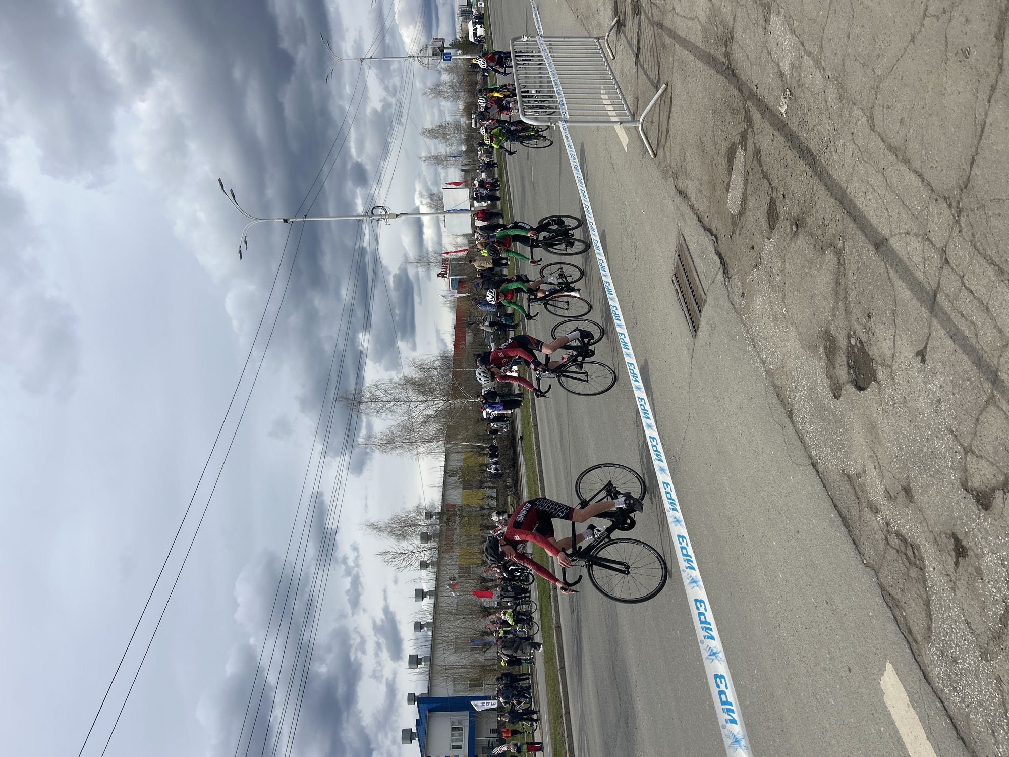 This vertically oriented photograph captures an outdoor road cycling event, likely a race or time trial, on a cloudy day in Izhevsk, Udmurtia, Russia. The scene features multiple cyclists in racing gear and helmets, positioned along a paved road marked with blue and white barrier tape.
In the foreground, several cyclists are visible, either actively riding or preparing to start. The lead cyclist, wearing a dark red and black jersey with "УДМУРТІЯ" (Udmurtia) text, is bent over his racing bicycle. Behind him, other cyclists in green, red, and black jerseys are also on their bikes. Spectators line the side of the road, separated from the course by metal barriers, observing the event.
The road surface is asphalt, showing signs of wear with cracks and patches. A storm drain is visible on the right side of the road. Overhead, streetlights and numerous utility wires stretch across the frame. The sky is heavily overcast with dark, voluminous clouds, suggesting cool or inclement weather. Bare, deciduous trees in the mid-ground indicate either early spring or late autumn.
In the background, various buildings are visible, including one with "NDE" written on a white sign and another displaying "УДМУРТІЯ" (Udmurtia). A red banner with "Ашан" (Auchan) text can also be seen. The blue and white barrier tape lining the race course prominently features the repeated text "ИРЗ" (IRZ), likely a local sponsor or organizer. The overall impression is one of an organized sporting event taking place in an urban setting under a dramatic, cloudy sky.