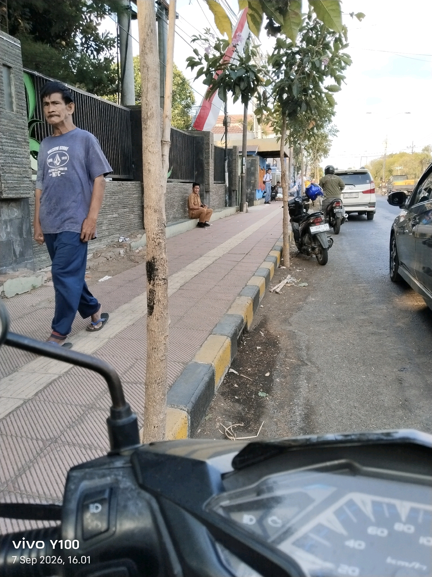 The image captures a street scene in Situbondo, Indonesia, on September 7, 2026, at 16:01. In the foreground, the handlebar and dashboard of a motorcycle are visible, indicating the photographer's perspective. To the left, an elderly man with short, dark hair and a mustache is walking on a sidewalk. He is wearing a blue t-shirt with a graphic and dark blue pants, and is shod in flip-flops. Behind him, a fence with vertical metal bars encloses an area with stone walls. Further down the sidewalk, a person in a light brown uniform is seated. The sidewalk is paved with textured tiles and bordered by a curb with yellow and gray stripes. A tree with a rough, gray trunk stands prominently on the sidewalk. The street adjacent to the sidewalk is paved with asphalt and shows signs of wear. Several vehicles are present, including a silver minivan parked on the right side of the road and a black car partially visible in the far right of the frame. A scooter is parked on the side of the road near the tree. In the background, trees and buildings are visible, with a partial view of an Indonesian flag. The sky is overcast.