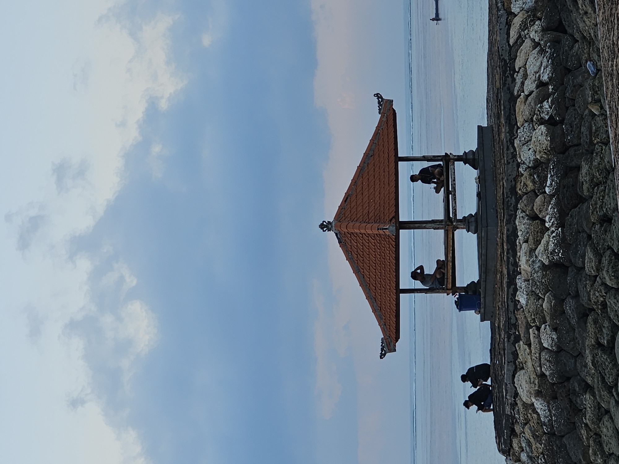 The image depicts a serene coastal scene at sunset, likely in Denpasar, Indonesia, given the architecture of the pavilion. In the foreground, a breakwater constructed of large, grey and white stones is visible, leading to a raised platform. On this platform, a traditional open-air pavilion with a terracotta-tiled roof stands prominently.
Within the pavilion, two individuals are seated. One is on the right side, looking towards the ocean, with his hand touching his head. Another person is on the left side of the pavilion, also seated. A blue plastic bin is placed near the left side of the pavilion.
Outside the pavilion, to its left, three figures are seated on the edge of the platform, facing the ocean. Two of them appear to be looking at their phones.
The background features a calm ocean stretching to the horizon, under a sky filled with soft, diffused clouds that suggest twilight. A small boat is visible in the far distance on the water. The overall atmosphere is tranquil and contemplative.