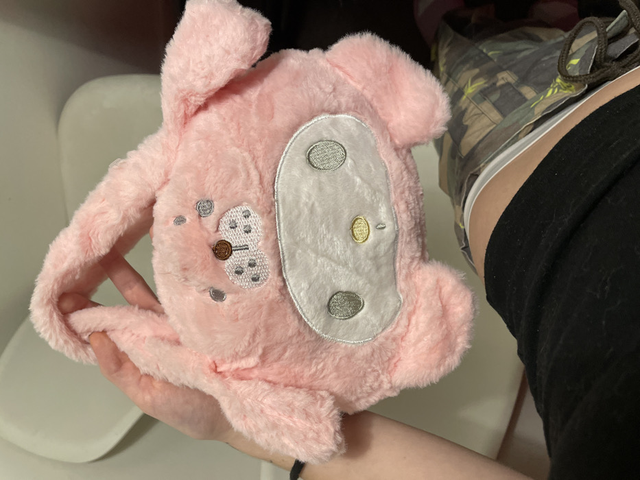 A pink, plush rabbit-like bag is held in a hand. The bag features a white, oval patch with three silver-embroidered shapes and a gold button-like feature. On the pink plush, a small butterfly shape is embroidered with silver dots and a brown center. The bag has long, floppy ears and is made of soft, fuzzy material. Black leggings and camouflage shoes are visible in the background.
