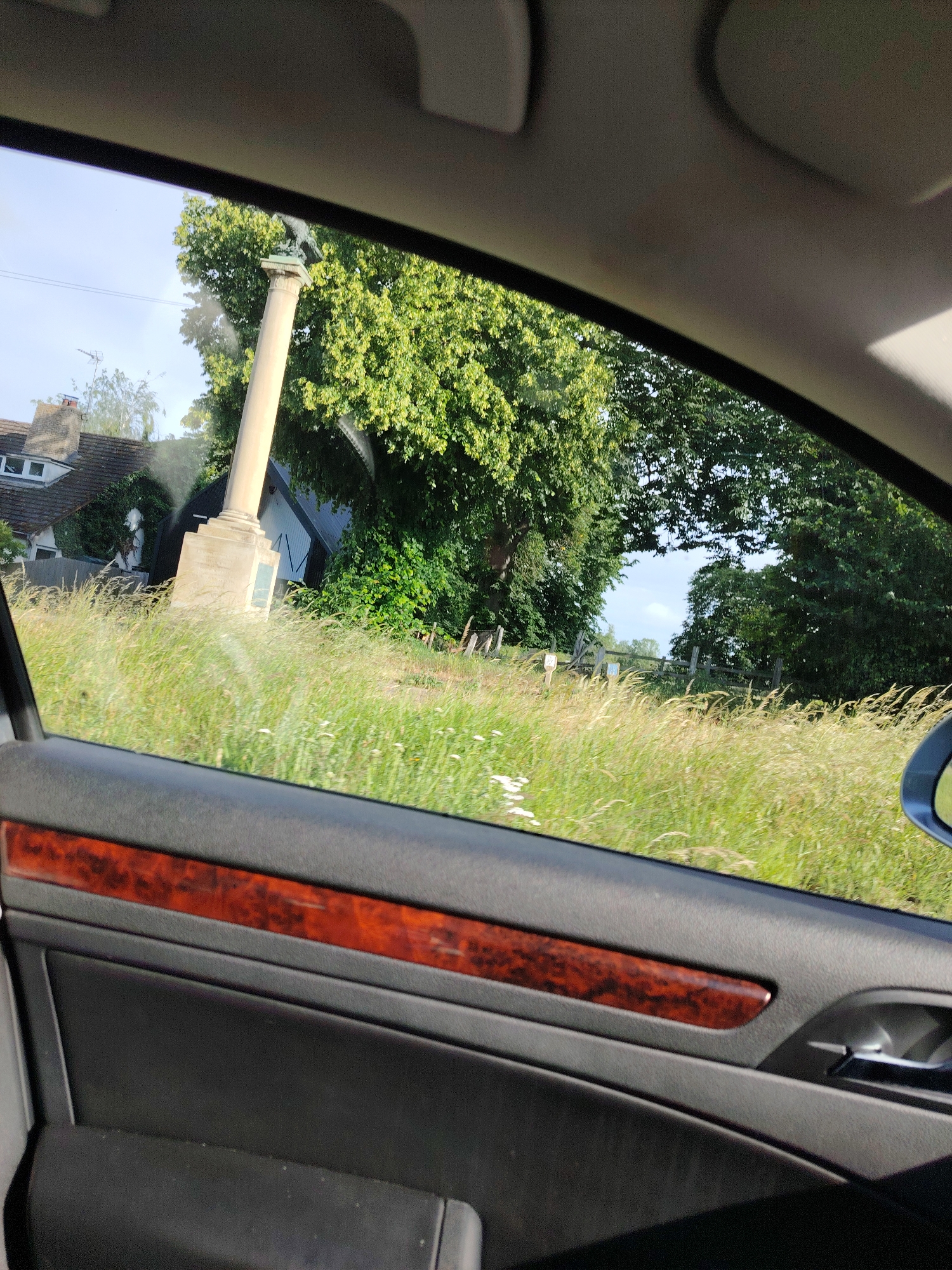 The image depicts a view from the interior of a vehicle, looking out through the passenger side window. The upper section of the image shows the car's grey headliner, while the lower section reveals a black door panel detailed with a reddish-brown, patterned wood trim and a silver-colored door handle.
Outside the vehicle, a rural landscape is visible under bright daylight. In the immediate foreground, tall, wild grasses with interspersed white wildflowers dominate the field of view. The grass exhibits slight motion blur, suggesting the vehicle is moving.
The central subject outside is a prominent, light-colored stone column, identified as the War Memorial located in Spaldwick, Huntingdonshire, United Kingdom. The column stands on a substantial pedestal base, which features an unreadable recessed panel. Surmounting the column is a dark-colored sculpture of an eagle with spread wings.
To the left of the memorial column, a residential building with a dark, pitched tile roof and dormer windows is partially visible. A white statue or figure is also present near the house, partially obscured by vegetation. Behind the column, a dark, barn-like structure with white vertical accents is discernible amidst green foliage. To the right of the scene, a rustic wooden fence line extends through dense green trees and undergrowth. The sky above is a clear light blue with minimal cloud cover. No individuals are present in the frame.
