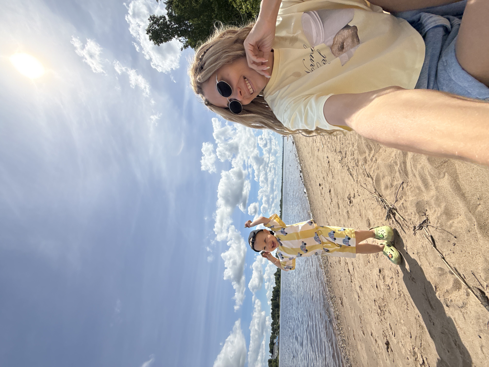 A high-angle, wide-angle selfie taken outdoors on a sunny day captures a woman and a young child on a sandy shore. The image is rotated, presenting a tilted perspective.
In the foreground, a woman with long blonde dreadlocks smiles broadly at the camera. She wears round, dark sunglasses, a light yellow t-shirt with a graphic of a white cup containing a donut, and blue shorts. Text partially visible on her shirt includes "energy." Her right hand is near her face, possibly adjusted for the selfie.
Further down the sandy incline, a young child, likely a boy, strikes an energetic pose with arms raised and a wide smile. He wears a yellow and white striped short-sleeved top and matching shorts, both decorated with cartoon car patterns. A dark cap and light green sandals complete his outfit.
The setting is a sandy beach alongside a calm body of water, likely a river or lake, extending into the background. A treeline is visible on the far bank. The sky is bright blue with scattered white, fluffy clouds, and the sun is prominently visible in the upper left corner, casting a bright glow. The overall mood is joyful, suggesting a pleasant day out in Rusanovo, Russia.