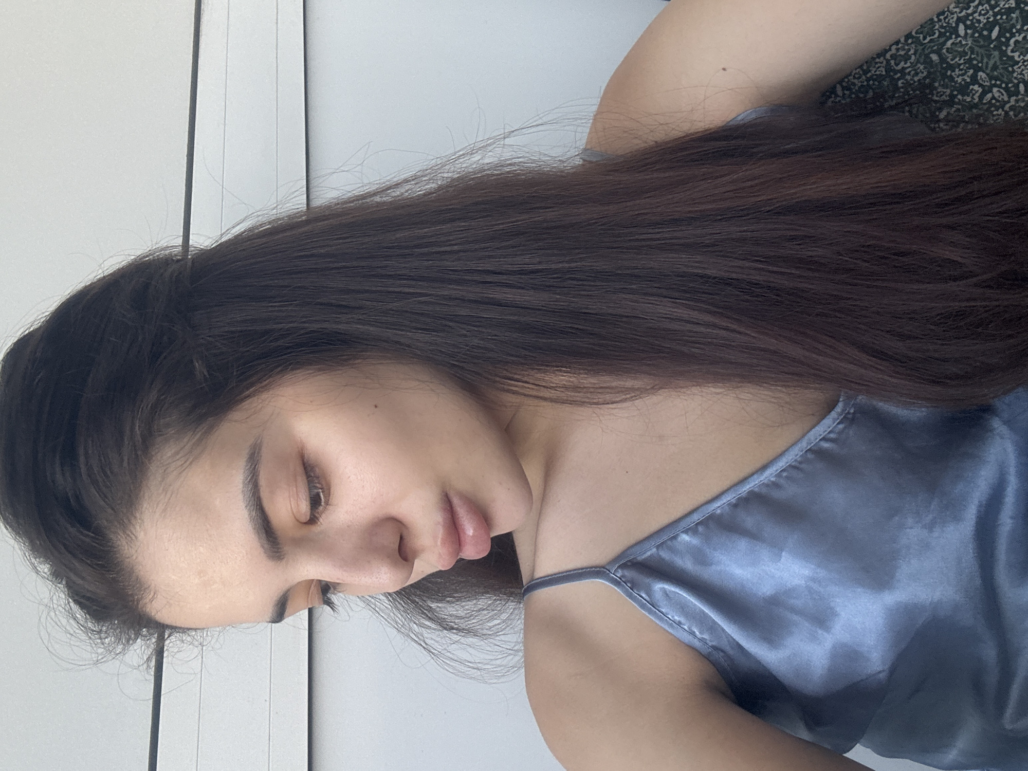 The image captures a young woman lying down in an indoor setting, likely a private room such as a bedroom. Her head is turned to the left side, and her eyes are cast downwards, giving her a calm and contemplative expression. She has long, dark brown hair, which is styled with some strands pulled back from her face, and the rest falling over her shoulder and arm. Some fine, wispy hairs are visible around her forehead and hairline.
She is wearing a light blue-grey top with thin straps, made of a smooth, reflective fabric that appears to be satin or silk. Her skin is clear, and her lips are slightly parted. In the background, there are light-colored, possibly white or light grey, vertical panels, which could be part of a wall, a wardrobe, or blinds. To the right of the frame, behind her shoulder, a dark fabric with a subtle, abstract pattern is visible, suggesting a bedsheet, pillow, or blanket.
The lighting is soft and bright, suggesting natural daylight. The overall atmosphere is one of relaxation and quiet introspection. No visible text is present in the image. While no specific architectural or environmental cues are visible to confirm the location, the image context indicates Plovdiv, Bulgaria.