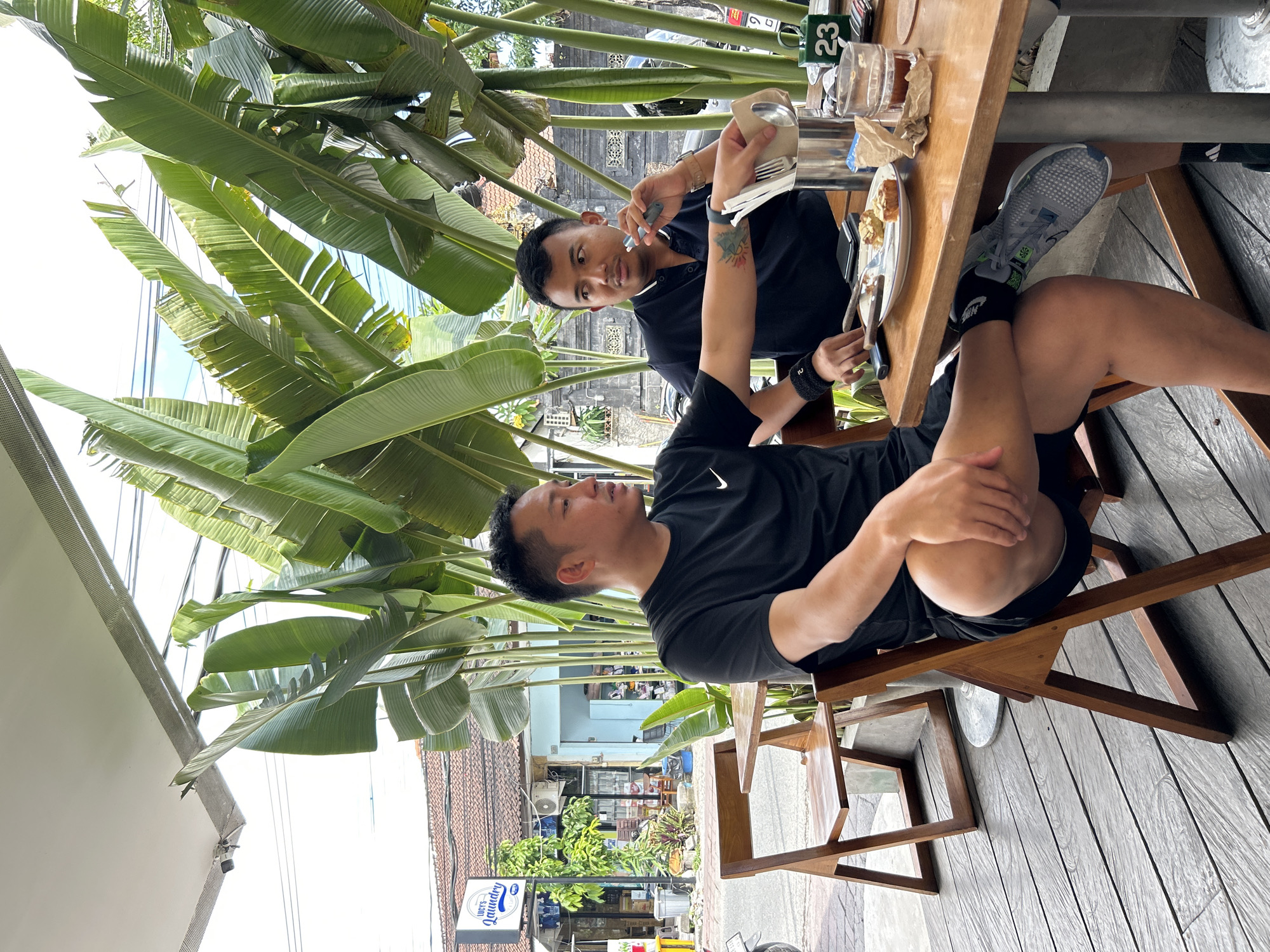 Two men are seated at a wooden table outdoors, with large green plants behind them. The man on the left, wearing a black t-shirt and black shorts, has his legs crossed and is looking to the side. The man on the right, wearing a black collared shirt, is holding a vaping device and looking towards the table. On the table, there are plates with food, cutlery in a metal container, and glasses. In the background, there is a sign that reads "LUCY'S Laundry" and buildings that appear to be shops or residences. The scene is set in a location that is likely a cafe or restaurant in North Kuta, Indonesia, given the presence of tropical foliage and the architecture.