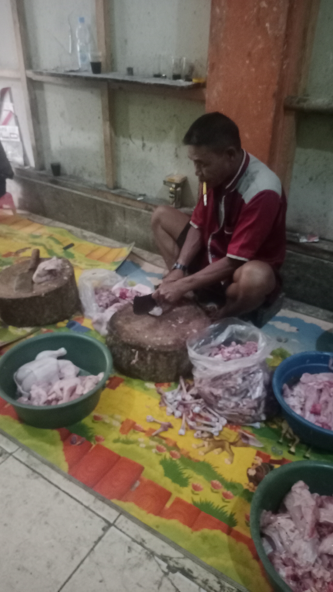 A man is sitting on the floor in a room with a concrete floor and walls, chopping meat on a wooden block. He is wearing a red and gray short-sleeved shirt and dark shorts. He is holding a large cleaver in his right hand and appears to be in the process of cutting pieces of meat. In front of him are several containers and bags filled with raw meat, including a green bowl containing a whole chicken and chopped chicken pieces, a large round wooden cutting board, a bag of chopped meat, and a blue bowl also containing chopped meat. To his left is another large wooden cutting board with a whole chicken on it. The floor is covered with a colorful mat with images of animals and flowers. In the background, there are shelves with bottles and glasses. The scene appears to be in a butcher shop or a food preparation area in Karyamandala, Indonesia.