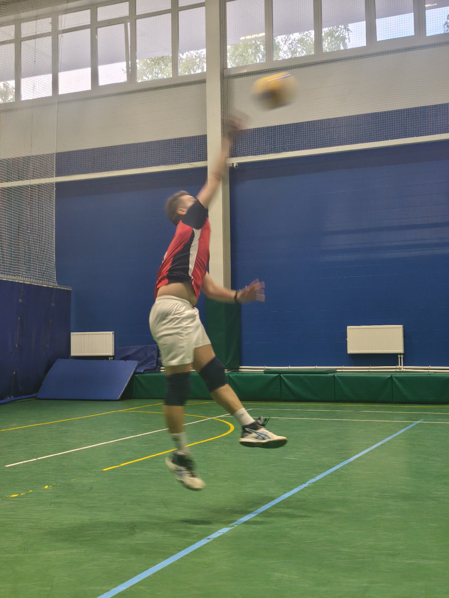 A man in a red and black jersey, white shorts, and knee pads is jumping in the air to hit a volleyball in an indoor gymnasium. The gym has a green floor with yellow, white, and blue lines. The walls are blue with a grid pattern near the top. There are radiators and a blue mat against one wall. A net is visible in the upper left corner. Windows are at the top of the wall. The image does not have enough information to identify a specific location in the city of Saint Petersburg, Russia.