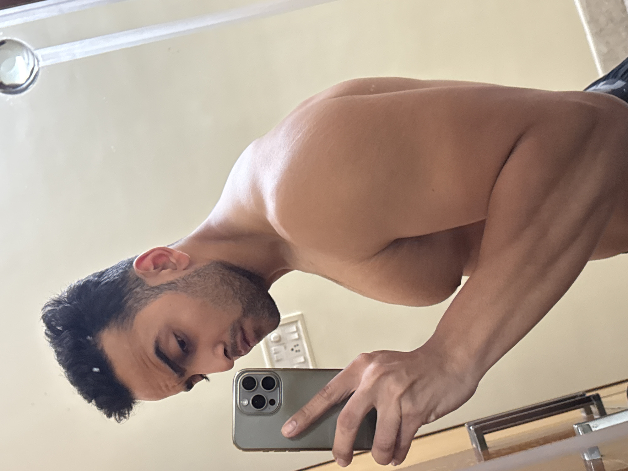 A shirtless man with a muscular build is taking a selfie in a mirror. He is holding a smartphone with a triple-lens camera in his right hand. His head is tilted, and he is looking at the phone screen. The background shows a beige wall with a light switch and a light fixture on the ceiling. The lower part of the mirror reflects a wooden surface and a chrome towel rack. There is no evidence to suggest this image was taken in Kolkata, India.