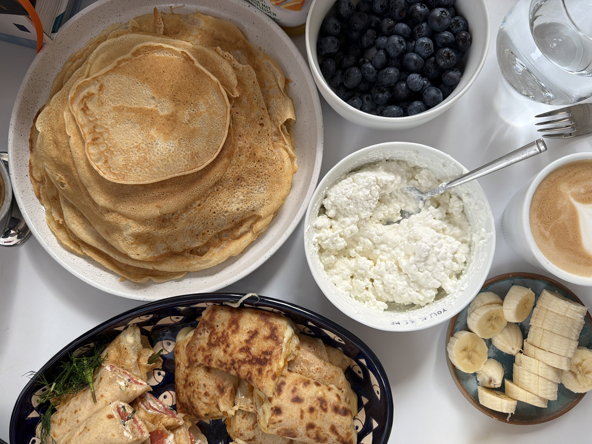 An overhead shot displays a breakfast spread on a white surface. In the foreground, a sliced banana rests on a small, blue-patterned plate. To the right, a white bowl is filled with fresh blueberries, and next to it, another white bowl contains a generous portion of cottage cheese, with a fork resting in it. A partially visible cup of coffee with latte art sits in the lower right corner, beside a glass of water and a silver fork. In the center, a large white platter is piled high with golden-brown crepes. To the left, a dark blue oval dish contains rolled crepes filled with what appears to be salmon and cream cheese, garnished with dill. The setting does not provide any context to determine its location in the city of Gangloff, Germany.