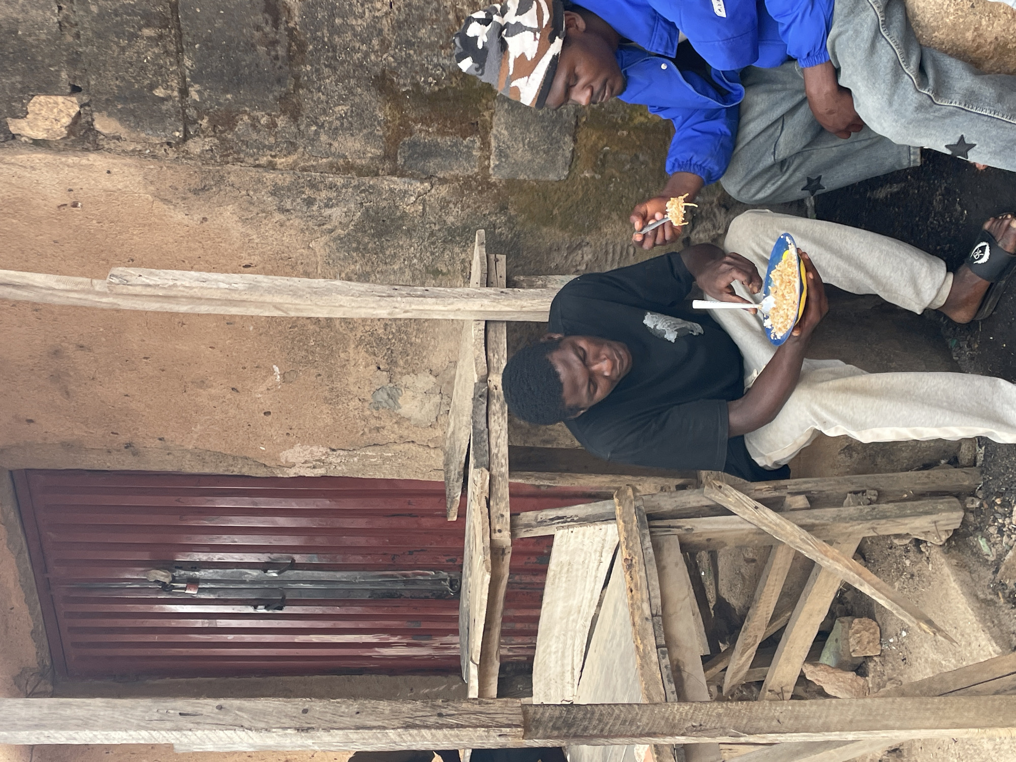 Two young men are eating from plates in an outdoor area in Jos, Nigeria. One man wears a black shirt and light-colored pants while the other wears a blue jacket, gray pants, and a patterned hat. Both are seated on the ground. The ground around them appears to be dirt and gravel. A red corrugated metal door is visible behind them, as well as a structure made of rough wooden boards. The setting appears to be somewhat impoverished.