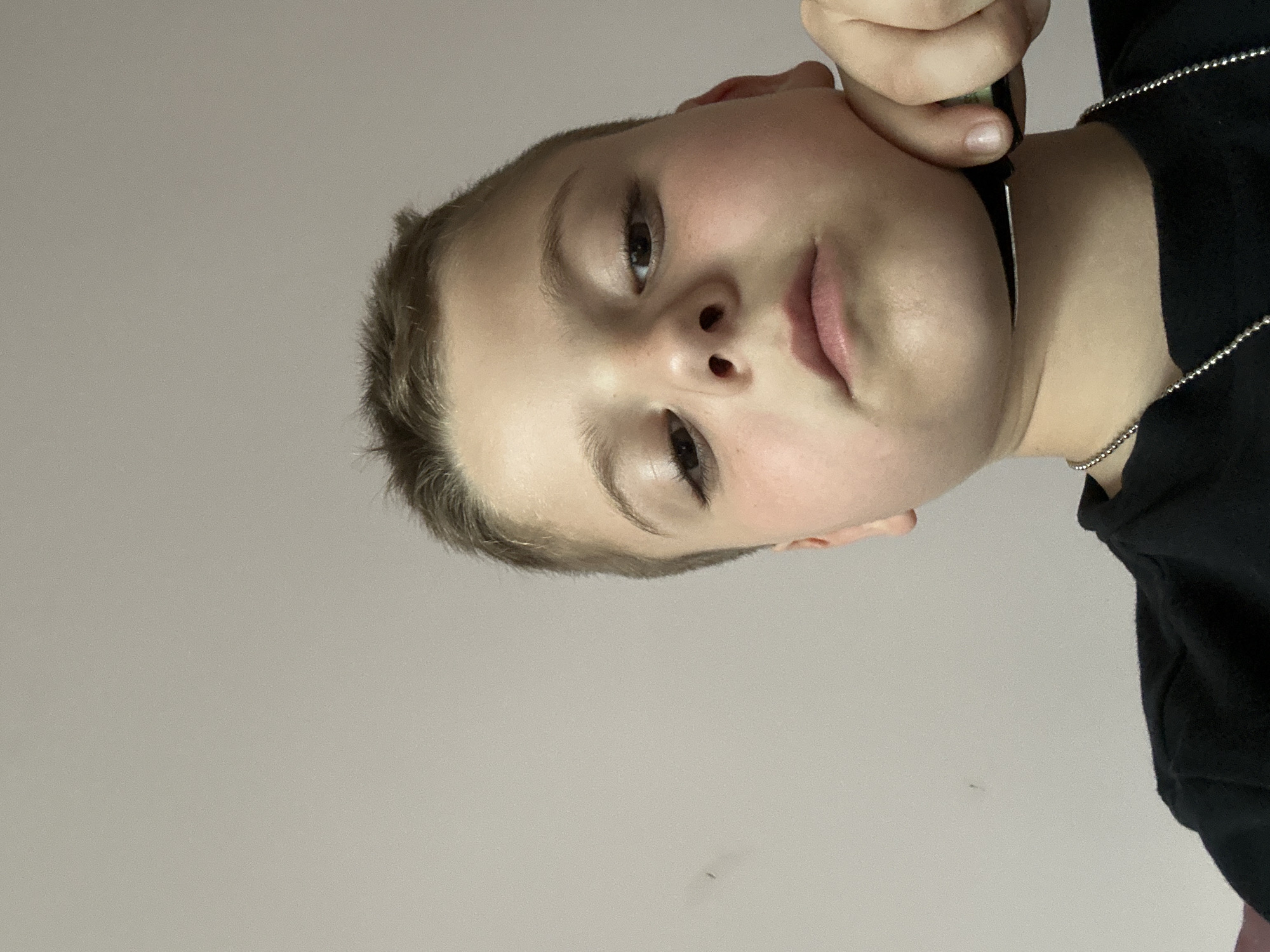 A close-up, slightly angled shot of a person with fair skin, light brown hair, and visible eyes, nose, and lips. The person is holding a black object near their face, possibly a pen or similar instrument. A black shirt or garment is visible on the right side of the image, and a silver chain is near the neck. The background is a plain light grey or white. There is no indication of location, city, or environment, and no interaction visible beyond the person holding an object near their face.