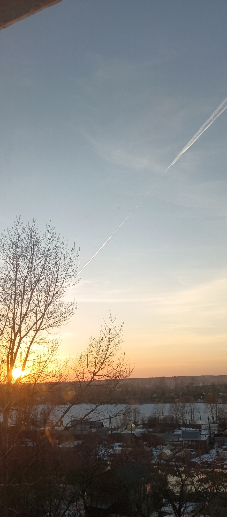 The photograph shows a cityscape at sunset. In the foreground, a bare tree silhouette is partially obscuring the sun, which emits a golden glow. In the background, there is a river or body of water bordered by trees. Beyond the river is an area consisting of buildings, possibly houses, interspersed with trees. Above the buildings and trees, the sky transitions from a lighter blue to orange and gold closer to the horizon. A contrail stretches across the upper part of the sky.