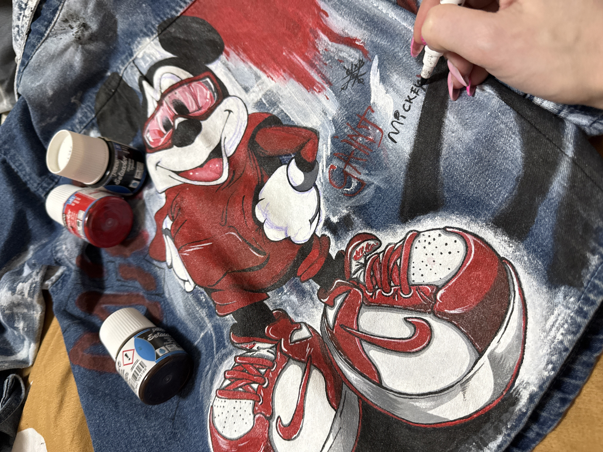 A partially painted denim jacket lies on a tan surface. The jacket features a large graphic of Mickey Mouse wearing sunglasses and a red jacket, with a pair of red and white sneakers. The design includes the word "Mickey" being written in black marker by a hand with long pink nails. Several small bottles of paint, including red and blue, are positioned near the artwork. The environment suggests an indoor workspace where the jacket is being customized. There is no evidence that the image was taken in the city of Chaikovsky, Russia.