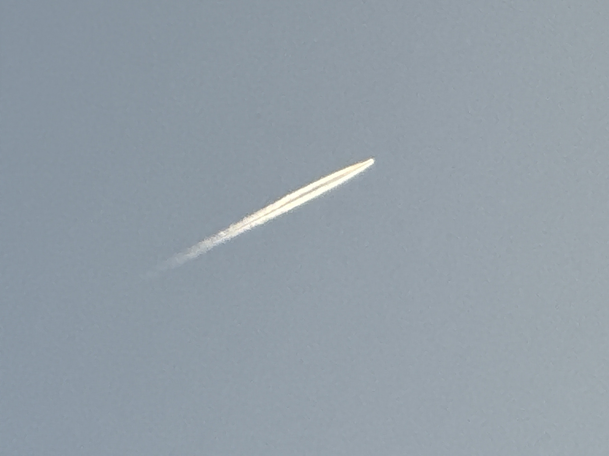 A faint contrail, likely from an aircraft, is visible in the pale blue sky above Budva, Montenegro. The contrail is thin and appears to be dissipating, with a brighter, more concentrated line at its center that tapers off into a wispy trail. The sky is overcast, and there is no indication of any buildings, landforms, or other objects in the frame.