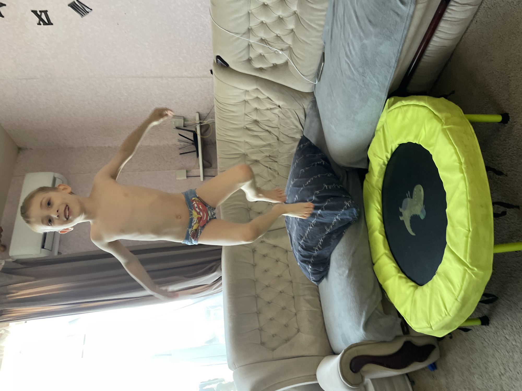 A young, shirtless boy is suspended mid-air, seemingly jumping. He is wearing patterned briefs. The boy is positioned above a beige, quilted sofa and a blue patterned pillow. Adjacent to the sofa is a small, round, green and black trampoline. The background reveals a room interior with a window, curtains, and wall decor. The location is not identifiable as Baranovka, Russia, based on the visual information provided.