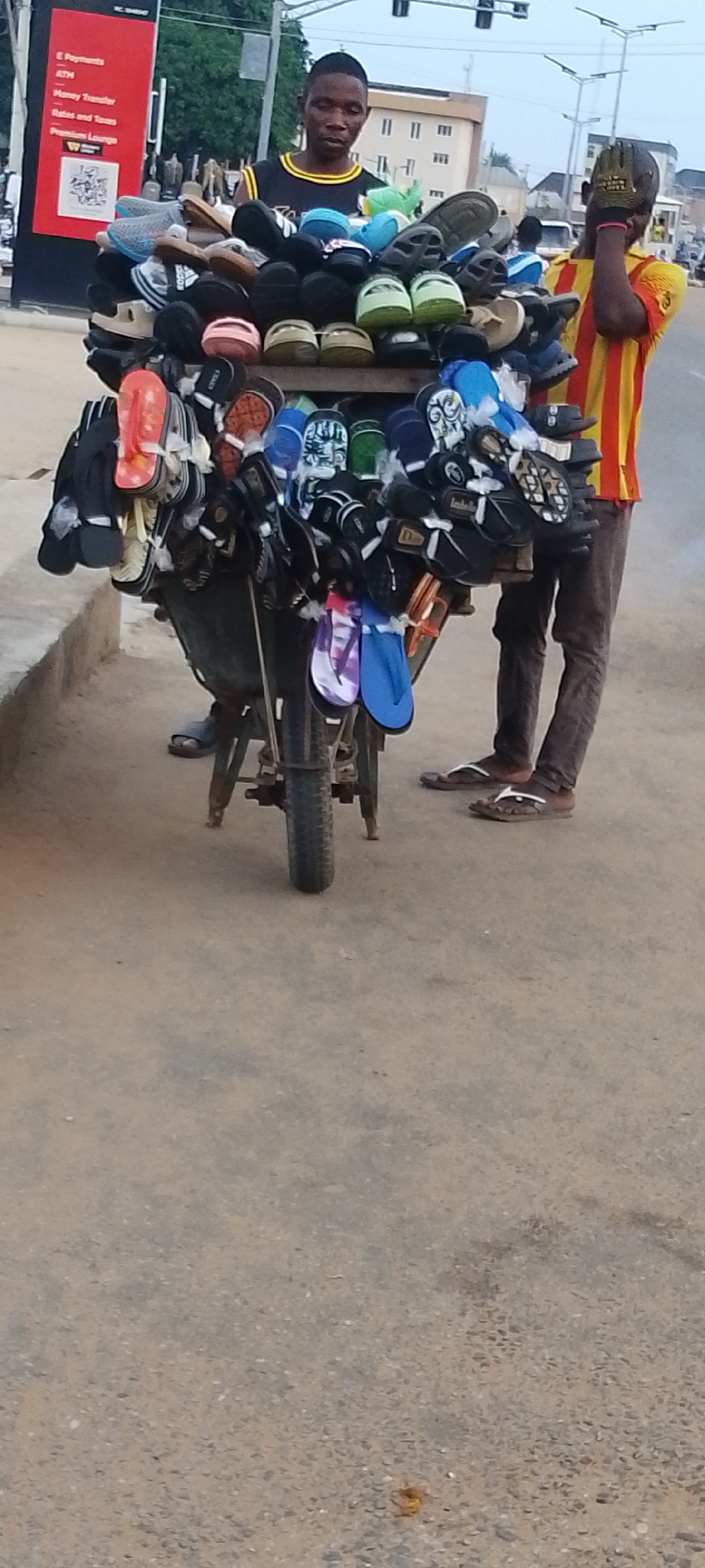 The image depicts two men in Lokoja, Nigeria, standing beside a wheelbarrow piled high with an assortment of sandals and flip-flops for sale. One man wears a black tank top with yellow trim and stands directly behind the wheelbarrow. The second man, on the right, wears a vertically striped yellow and red shirt and dark trousers and has a hand covering his face. Both men wear sandals. The wheelbarrow is positioned on the side of a road, and a curb is visible to the left. A sign with red background and white text is visible in the background on the left, and a building is seen across the road. Traffic lights are also visible in the background. The environment appears to be a busy street.