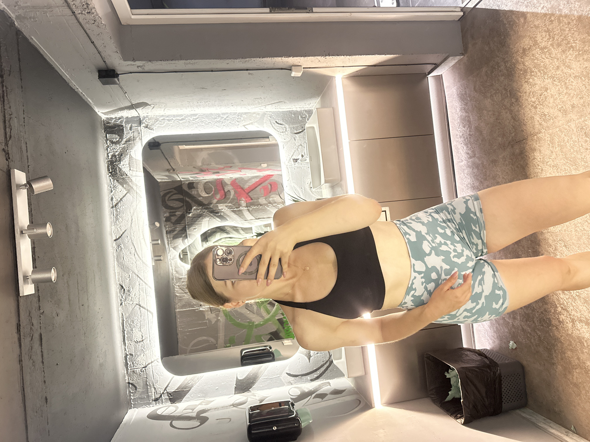 A woman is taking a mirror selfie in what appears to be a public restroom or changing area. She is wearing a black sports bra and light blue camouflage-patterned shorts. She is holding a smartphone in her right hand, capturing her reflection. The environment includes a large mirror with graffiti visible on the wall behind it, a white sink, lighting fixtures, a trash can, and a tiled floor. The background suggests an urban, possibly industrial or recreational setting. The specific location in Ternopil, Ukraine is not discernible from the image content.