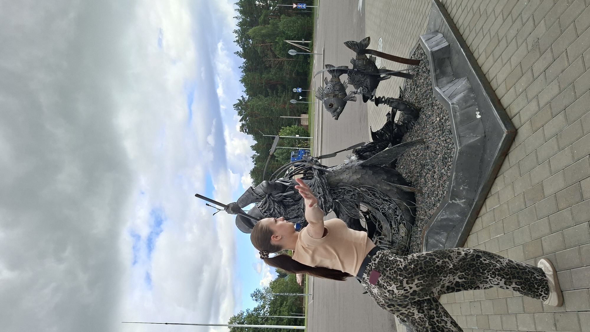 A young woman with a ponytail, wearing a light brown t-shirt and leopard-print pants, is posing in front of a metal sculpture. The sculpture depicts a figure, possibly a warrior, engaged in battle with fish. The woman is in the foreground, with her arms outstretched as if mimicking the sculpture's action or performing a dance. Behind her, the sculpture is situated on a paved area, surrounded by gravel. In the background, there is a road, trees, and a cloudy sky. The scene appears to be outdoors in a public space.