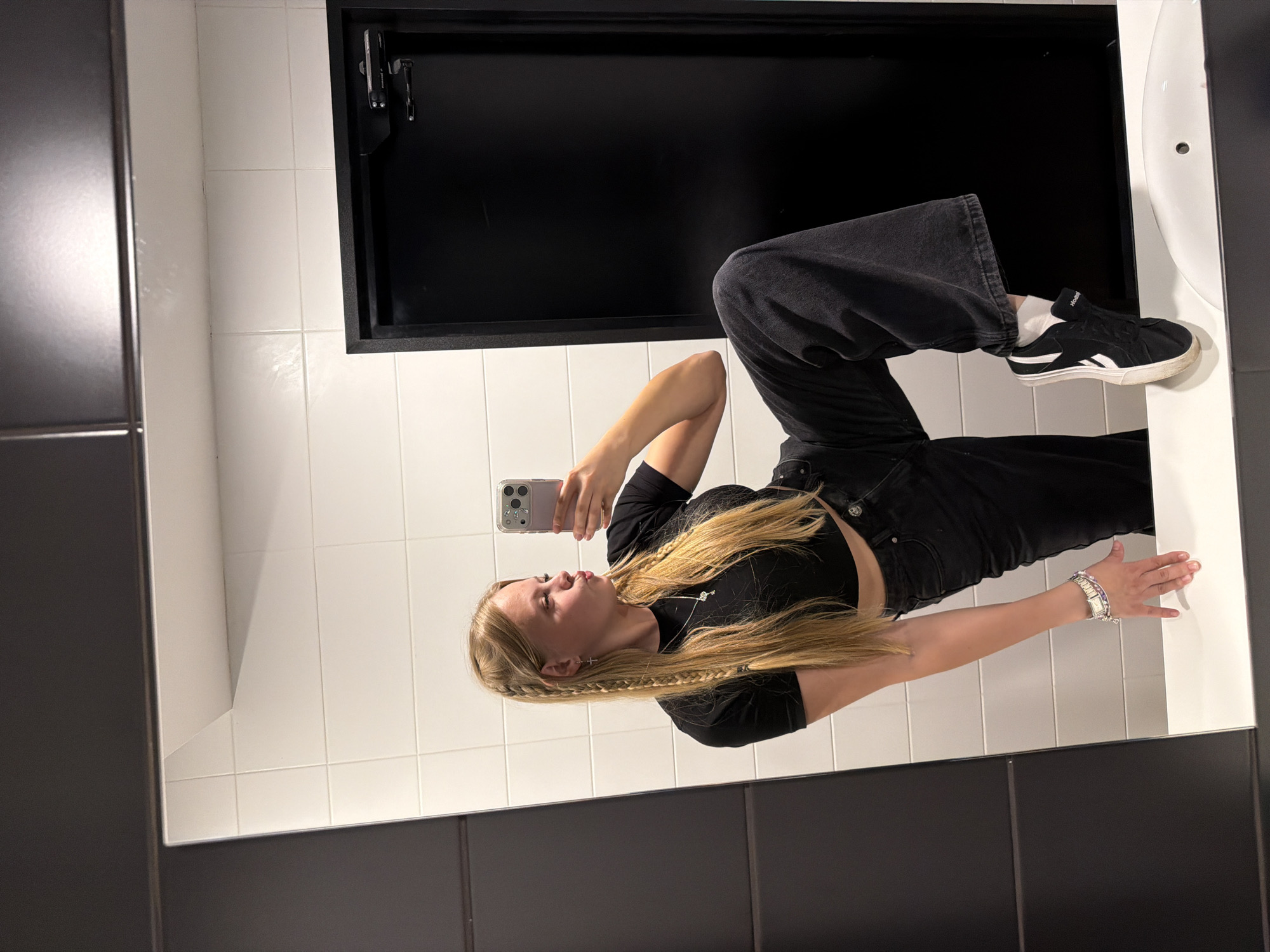 A young blonde woman is taking a mirror selfie in a restroom. She is wearing a black crop top, black wide-leg jeans, white socks, and black and white sneakers. Her hair is styled in two braids with some loose strands framing her face. She is pouting her lips and holding a pink smartphone in her right hand. Her left hand is resting on the counter. The restroom features white tiled walls and a black door. The overall impression is of a casual, stylish pose. The location is not determinable from the image.