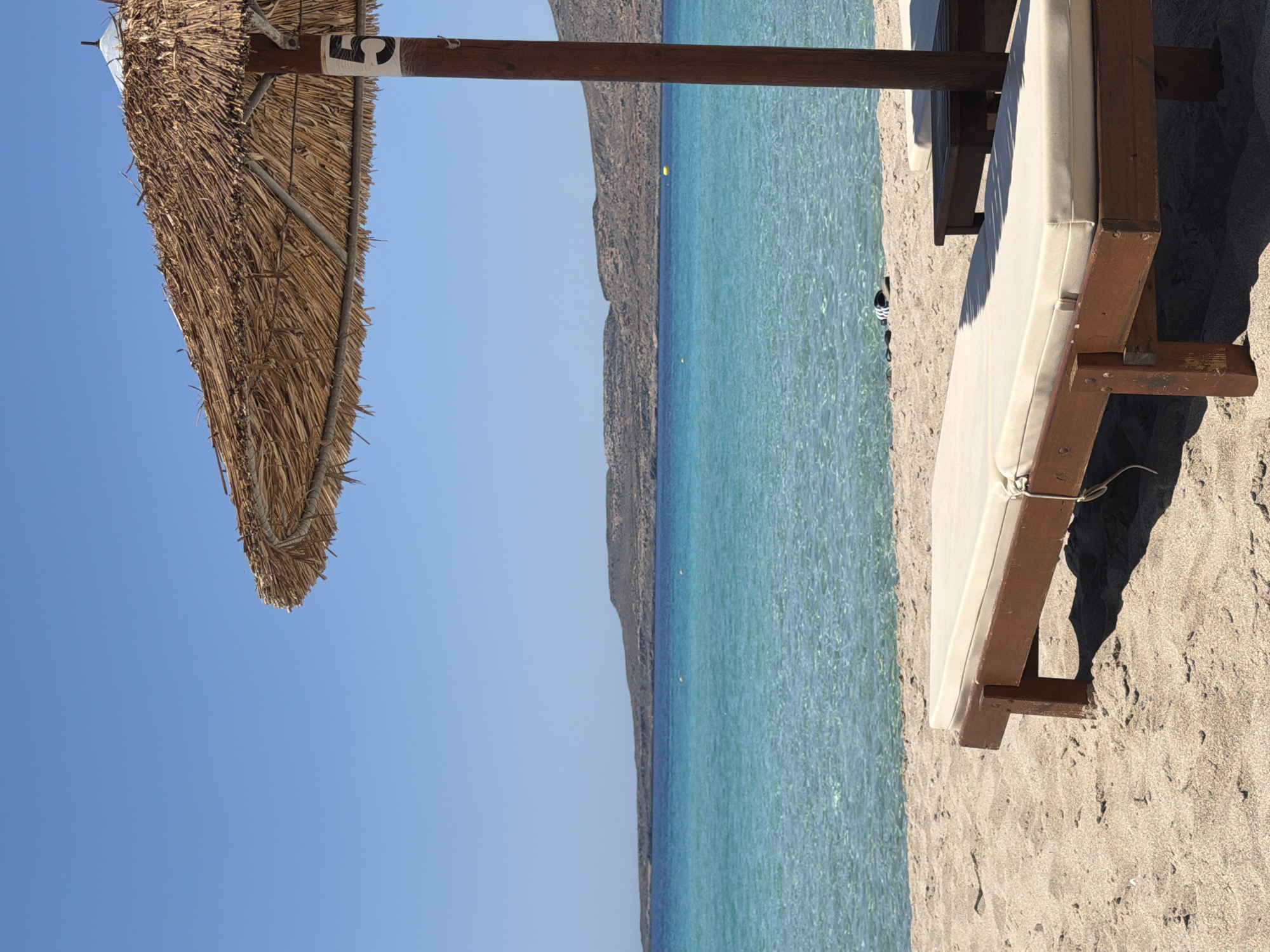 This image depicts a serene beach scene in an unknown location in Greece. A light-colored sunbed with a padded cushion rests on the sandy shore in the foreground. Behind it, another dark wooden table-like structure, possibly a smaller side table, is partially visible. A thatched umbrella, supported by a dark wooden pole with the number "5" marked on it, provides shade for the sunbed. The crystal-clear turquoise water of the Aegean Sea gently laps the shore, with a distant, low-lying, arid island forming the horizon. The sky is a clear, cloudless blue. There are no people visible in the image, emphasizing the tranquility of the setting.
