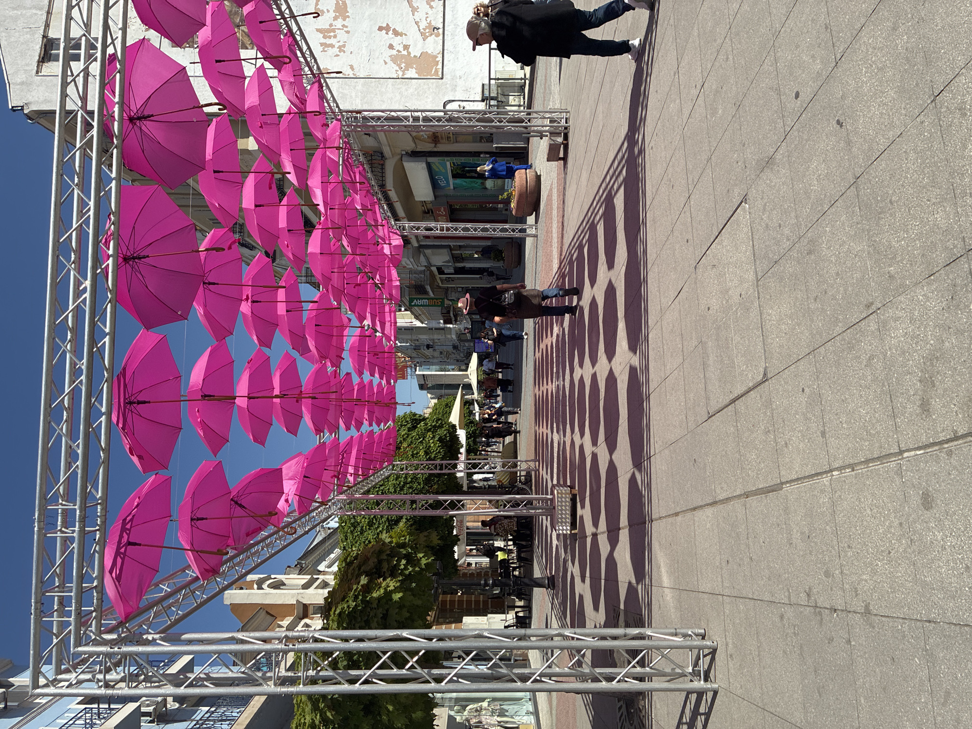 The image shows a pedestrian street in Plovdiv, Bulgaria, lined with pink umbrellas suspended from a metal scaffolding structure. The umbrellas create a canopy overhead, casting shadows on the tiled pavement. People are walking along the street, and there are businesses and restaurants visible in the background. The sky is blue and clear.