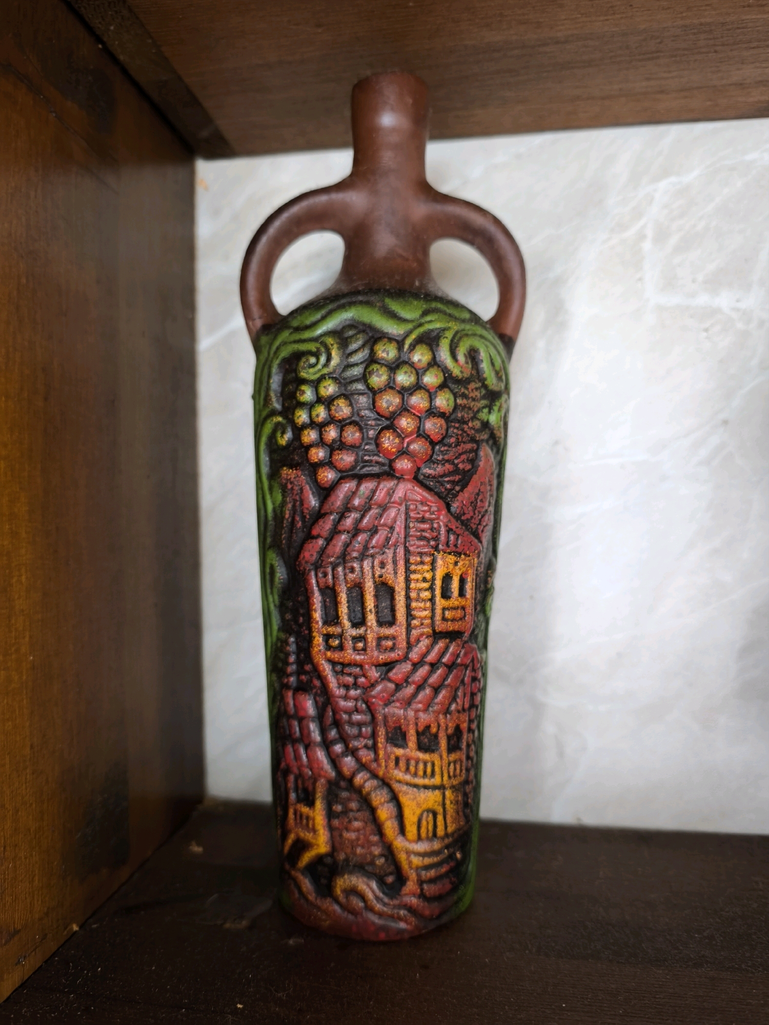 A clay bottle with carved relief decoration sits on a dark wooden shelf. The bottle has a narrow neck and two looped handles near the top. The relief depicts a scene with grapevines at the top, followed by buildings that appear to be houses with red tiled roofs and yellow windows. The carving is painted in various colors, including green, red, yellow, and brown. The background behind the bottle is a light-colored marbled surface. To the left of the bottle is a dark wooden shelf. The image does not depict the city of Vyshegorod, Russia.