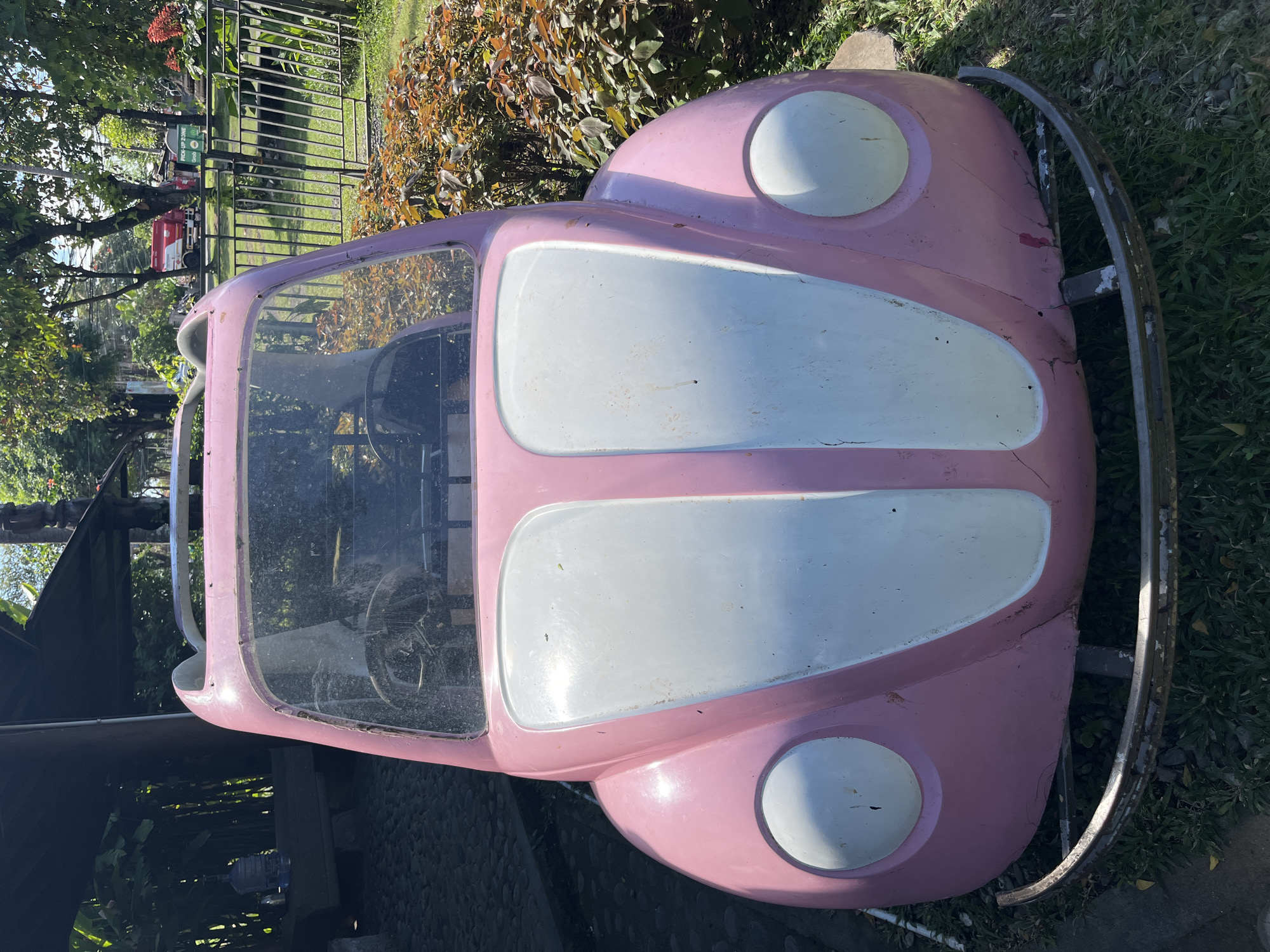 A pink toy car resembling a classic Volkswagen Beetle is positioned outdoors, likely in a public space. The car is bright pink with white accents on the hood and white circular headlights. It appears to be weathered, with some visible paint chips and cracks. The interior of the car, visible through the windshield, contains two steering wheels.
The car is situated on a patch of grass, with a metal bar or railing in front of it. Behind the car, there's a variety of foliage, including bushes with reddish-brown leaves and taller trees with green canopies. A dark-colored, possibly stone or paved, area is visible to the left of the car, with a wooden bench and a large blue water jug on it. In the background, a metal fence runs along the perimeter of the area, beyond which more greenery and what appears to be a building with a red and white truck are visible. The overall setting suggests an outdoor recreational area, possibly a park or a play zone. Based on the context of the previous images and the characteristic architecture and greenery, this location is identified as the Lapangan Puputan Badung in Denpasar, Indonesia.