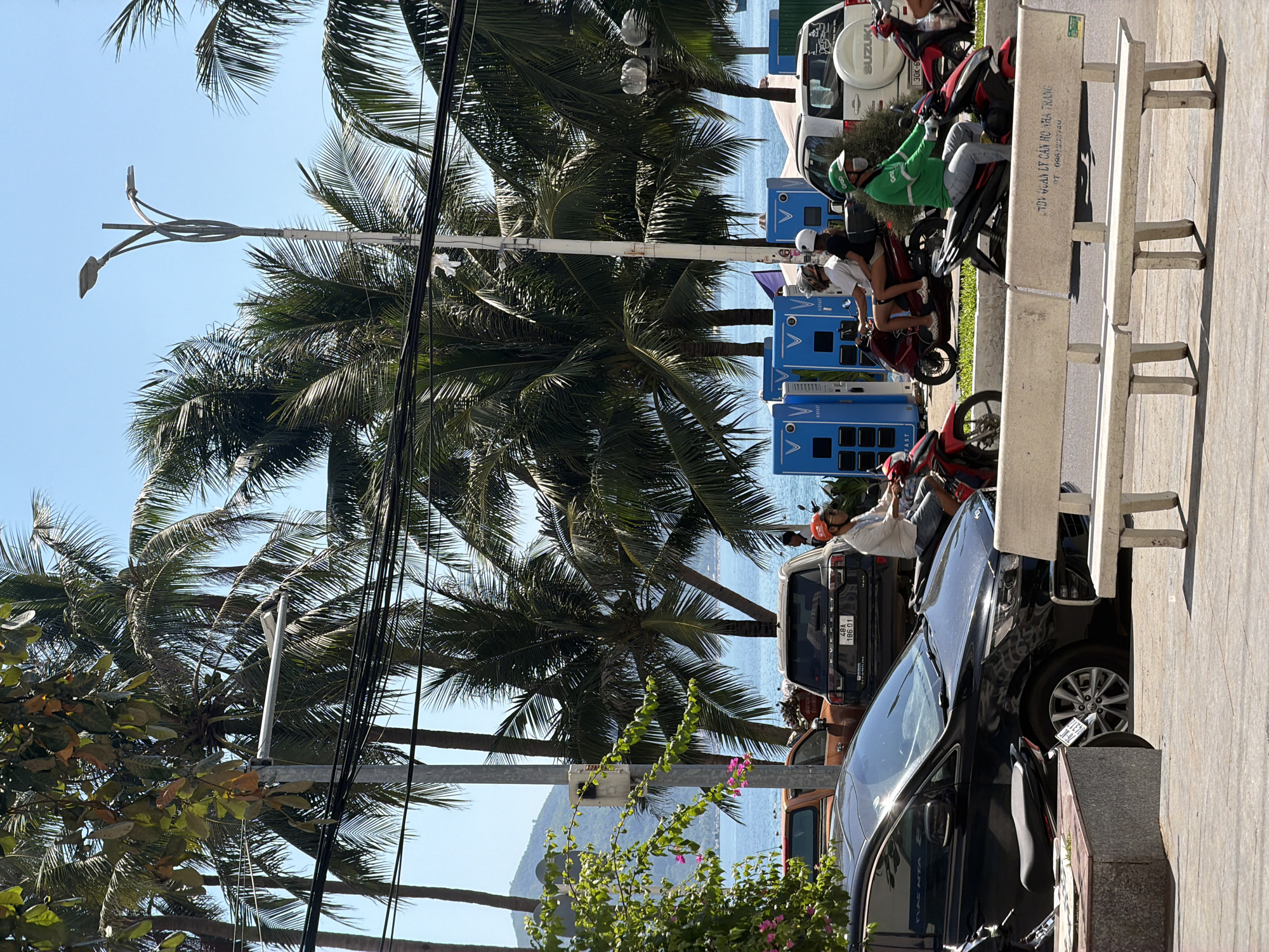 In Bac Nha Trang Ward, Vietnam, a sunlit street scene unfolds with a focus on transportation and urban infrastructure against a backdrop of palm trees and the ocean. A black Kia Carnival minivan, a common sight in Vietnam, is parked on the left side of the frame. To its right, a dark-colored SUV with a license plate reading "48A 186 01" is also visible. A white Suzuki SUV is positioned further back, on the right.
Several individuals are navigating the street on motorcycles. A person in a white shirt and helmet rides a red motorcycle, while another, also in a white helmet, is a passenger. A delivery driver, wearing a green jacket and a face mask, rides a black and red motorcycle, likely for a service like Grab, a popular ride-hailing app in Southeast Asia. The street is lined with palm trees, their fronds casting shadows on the pavement. In the background, the blue ocean is visible, with a few boats on the horizon. Electric vehicle charging stations, labeled "VinFast," are also present, indicating the growing adoption of electric mobility in the region. A concrete bench sits in the foreground, suggesting a public seating area. The overall impression is one of a typical day in a coastal Vietnamese city, characterized by a mix of traditional and modern elements.