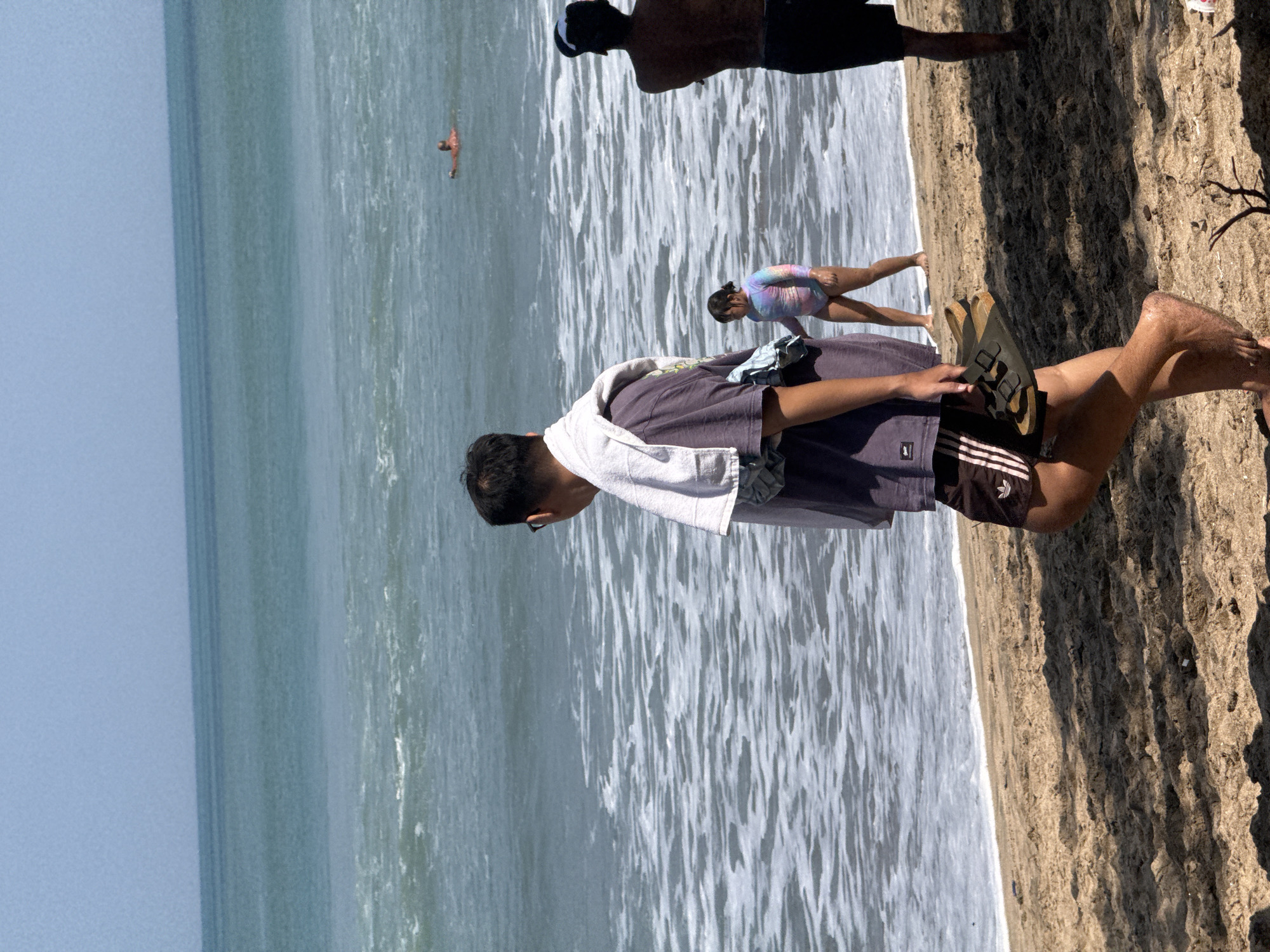The image depicts a beachfront scene in Kuta, Indonesia, on a sunny day with a clear sky and calm ocean conditions. In the foreground, a man wearing glasses and a purple t-shirt with a towel draped over his shoulders is walking away from the viewer, carrying a pair of sandals. He is wearing dark shorts with a white stripe. To his right, a young girl in a colorful swimsuit is walking towards the water. Further to the right and slightly behind, another person is standing with their back to the camera. In the background, a person is swimming in the ocean. The sandy beach is visible in the foreground, with gentle waves lapping at the shore. The overall atmosphere is one of a relaxed day at the beach.