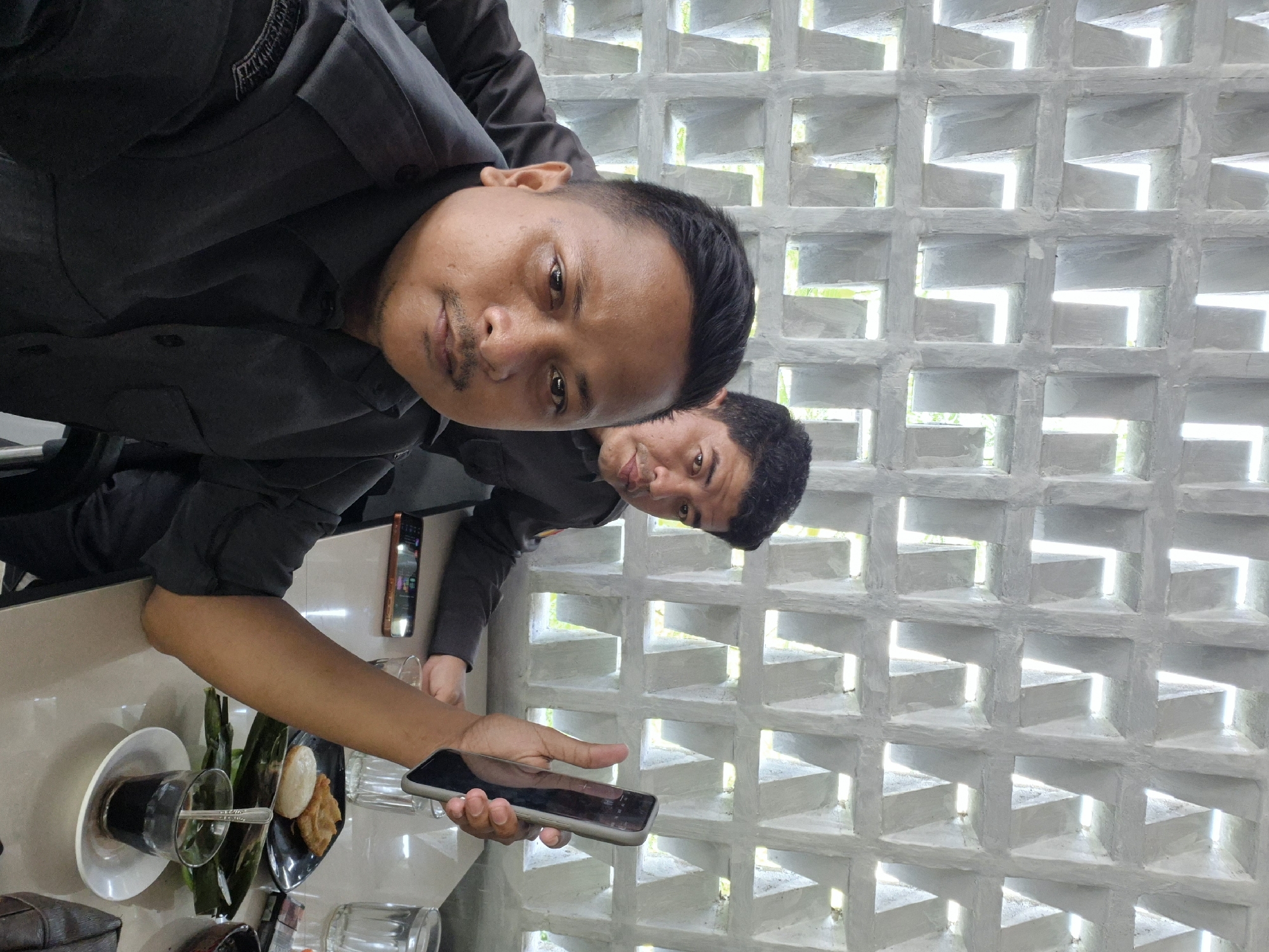 The image, taken in Lhokseumawe, Indonesia, captures two men in a casual indoor setting, likely a cafe or restaurant, under natural daylight. The photograph is oriented vertically, with both subjects facing the camera.
In the foreground, a man with short dark hair and a mustache dominates the left side of the frame. He wears a dark grey or black collared shirt, potentially a uniform given an unreadable patch on his right shoulder. He holds a dark-colored smartphone in his right hand, partially obscuring his face, and gazes directly at the viewer with a neutral to slightly smiling expression. His left arm rests on a light-colored table.
Behind and to his right, a second man, similarly dressed with short dark hair, is partially visible, also looking towards the camera with a subtle smile.
The table in front of them is laden with various items: a glass of dark liquid (likely coffee) resting on a white saucer with a spoon, another empty glass, and a black plate holding what appear to be local snacks—possibly fried items or cakes—garnished with green leaves, suggestive of banana leaves used for serving. A lighter and possibly a cigarette pack are also visible near the foreground man's left hand. Another smartphone with a glowing screen is positioned further back on the table.
The background features a prominent light grey or white concrete wall with a distinctive grid pattern of square, recessed openings. These openings allow bright, natural light to stream in, indicating it is daytime and possibly revealing an outdoor area beyond. The overall scene suggests a relaxed social gathering.