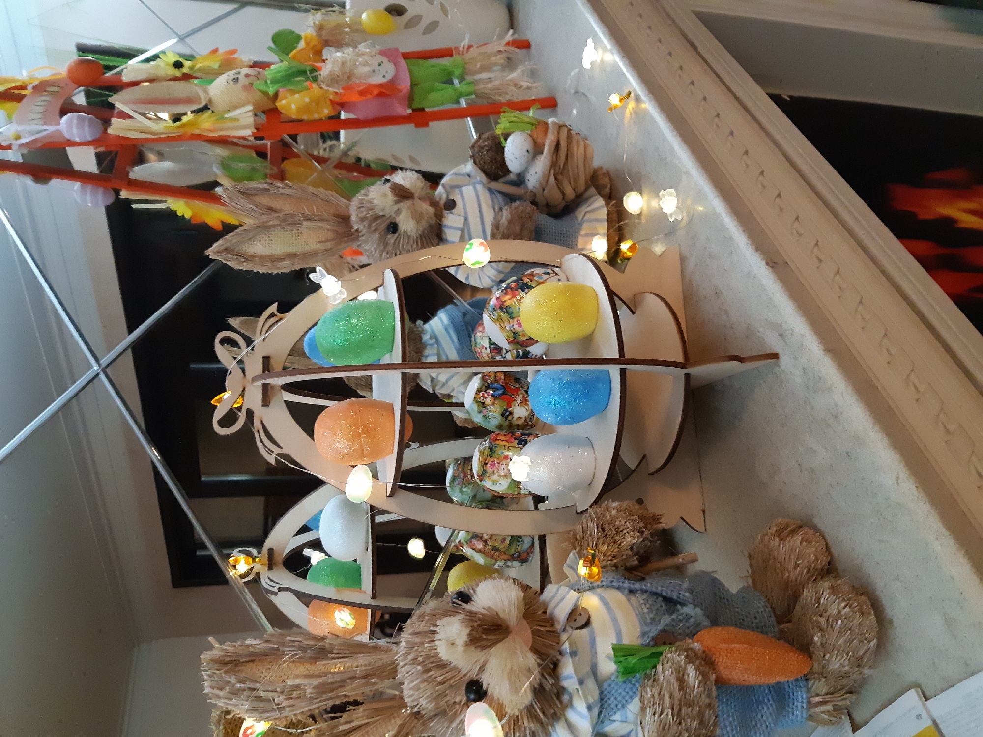 The image shows a decorative Easter display, likely indoors, in Plovdiv, Bulgaria. The display includes a wooden Easter egg holder filled with colorful eggs, two stuffed bunnies wearing striped clothing and holding carrots, and a tiered stand filled with various Easter-themed decorations. String lights with flower-shaped bulbs are woven through the display. The background is a kitchen counter with a patterned backsplash and a mirror reflecting the scene.