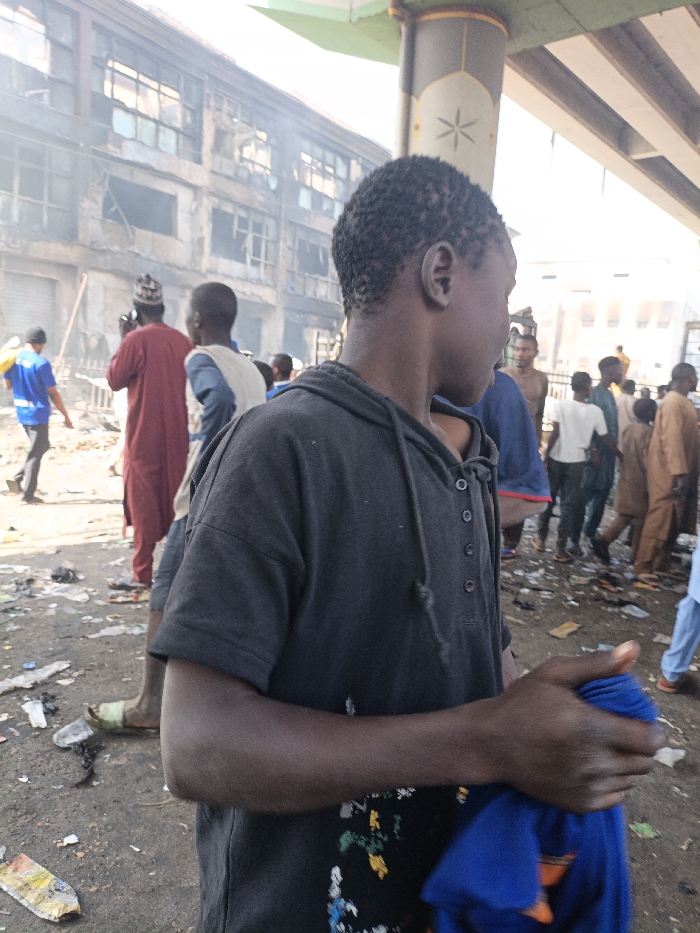 The scene depicts an outdoor urban environment in Fagge C, Nigeria, with a visible aftermath of an incident. In the immediate foreground, a young, dark-skinned male subject, estimated to be a teenager or young adult, stands with his back largely to the viewer, facing toward the right side of the frame. He wears a dark grey short-sleeved hooded shirt with a four-button placket and drawstrings; a faint pattern is present on the lower front. His right hand holds a folded piece of blue fabric.
Behind the foreground subject and to his left, a group of approximately 15-20 individuals, predominantly male, are gathered. One man in a red traditional garment and a dark cap is visible from the back, appearing to hold a device (possibly a phone or camera) toward a damaged building. Another man is partially visible in a light-colored vest over a blue shirt. Other individuals are seen standing or walking, dressed in various casual and traditional attires.
The background features a heavily damaged multi-story building, showing numerous broken windows, exposed interiors, and extensive charring or smoke discoloration on its facade. White smoke or dust emanates from various sections of this structure. To the right, a large concrete pillar, decorated with a star-like motif, supports an elevated concrete structure or bridge that extends across the upper right portion of the frame. The ground beneath the individuals is unpaved and covered with debris, including discarded plastic items, wrappers, and general refuse. The lighting suggests daylight conditions.