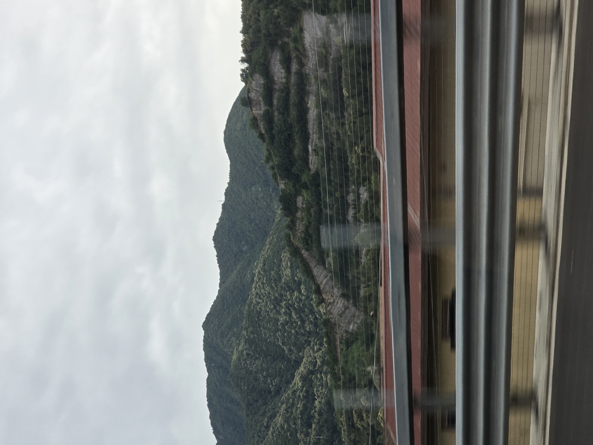 The image captures a scenic view of rolling, tree-covered mountains under a cloudy sky, likely taken from a moving vehicle in the vicinity of Baronissi, Italy. In the foreground, a highway guardrail is visible, with the blur of motion indicating the perspective of a traveler. Behind the guardrail, a building with a reddish roof and yellow siding is partially obscured. The mountains dominate the upper portion of the frame, displaying lush vegetation and visible terracing on some slopes, suggesting human cultivation or landscaping. The overall impression is one of a natural landscape viewed from a transportation route.