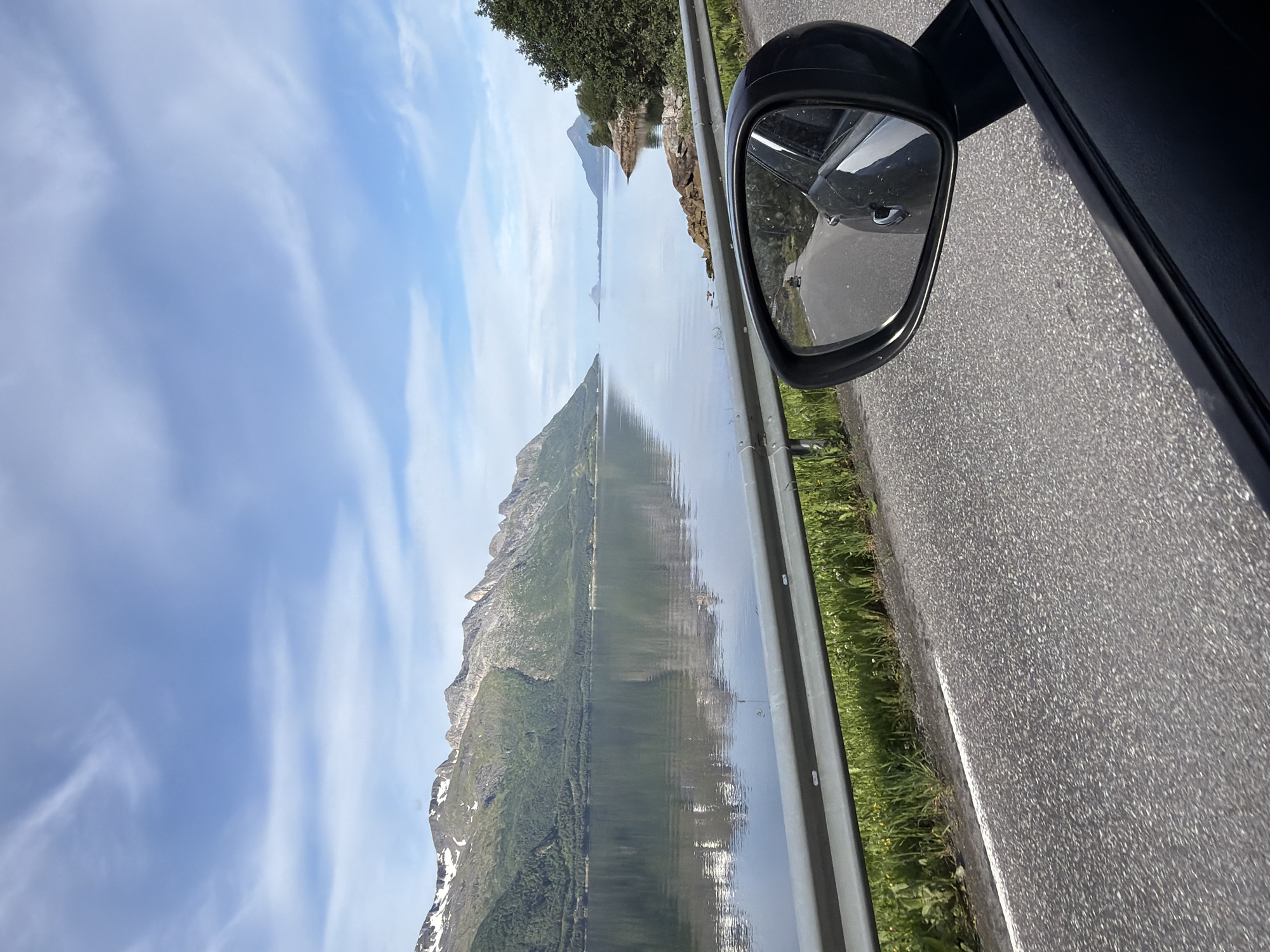 The image depicts a scenic view from a car traveling along a road near Neverdal, Norway. The foreground shows the asphalt road and a portion of the car's interior, including the side mirror which reflects the road behind the vehicle. A silver guardrail runs along the side of the road, separating it from a grassy verge. In the mid-ground, a calm body of water, likely a fjord, stretches out, reflecting the sky and the mountainous landscape. The mountains in the background are covered in green vegetation at the lower slopes and have patches of snow or white rock near their peaks. The sky is blue with scattered white clouds. The overall impression is of a serene, natural landscape viewed from a moving vehicle.