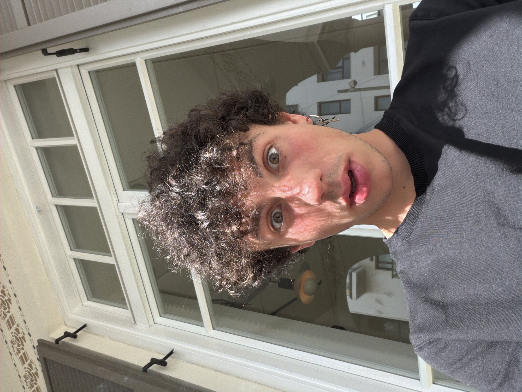 A headshot of a man with curly, dark hair is framed by a white-framed window. The man is fair-skinned with wide eyes and a surprised expression. He wears a dark-colored top. Reflected in the window are buildings and structures typical of an urban environment. The window hardware suggests an older architectural style. The photograph was taken from an upward angle, showcasing the city view. Considering the architectural style and urban density reflected in the window, the location is likely Tel-Aviv, Israel.