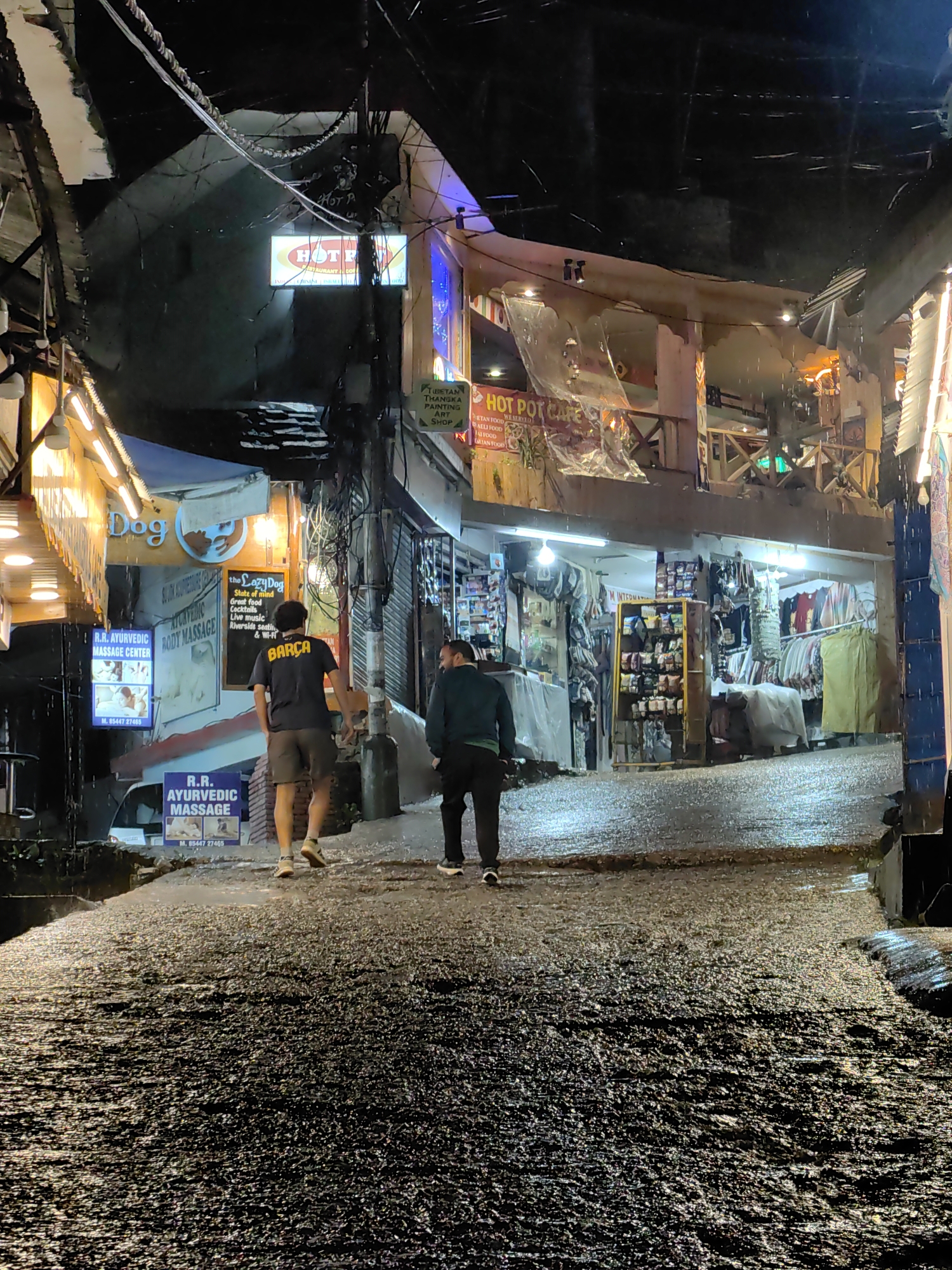 The image captures a wet, uphill street in Manali, India at night. Two men walk away from the camera, their figures silhouetted against the illuminated storefronts. The street is slick with water, reflecting the colorful lights of the shops. One man wears a black t-shirt with "Barça" printed on it, and the other wears a dark jacket.
On the left side of the street, a shop with a sign that reads "Dog" is visible, along with advertisements for "R.R. Ayurvedic Massage Center." Further up the street, a sign for "HOT POT CAFE" is illuminated, with its menu listing Tibetan, Indian, and Italian food.
The buildings lining the street appear to be a mix of shops and eateries, with goods displayed on shelves and hanging from racks. The scene is characterized by the wet conditions, the nighttime lighting, and the commercial activity of the street.