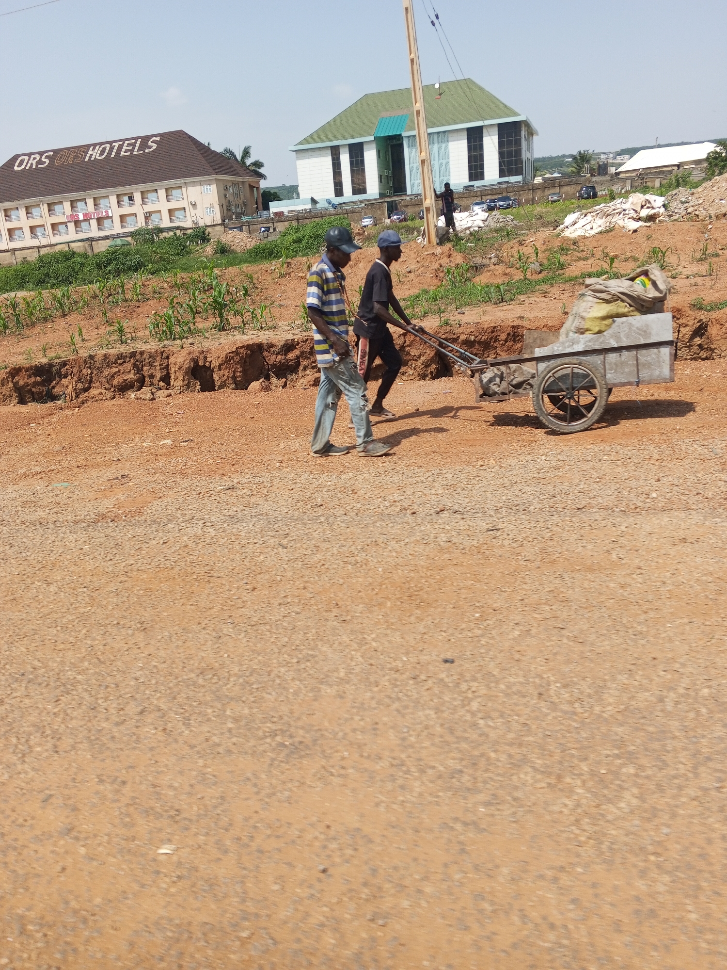 Two men are working on a dirt road in Abuja, Nigeria. One man, wearing a blue cap, striped shirt, and jeans, walks behind a two-wheeled cart. The other man, wearing a black shirt and dark pants, pushes the cart. The cart is filled with debris, possibly construction materials. In the background, there are buildings, including a hotel with "ORS ORS HOTELS" signage, and a light blue and green building with a green roof. The environment appears to be under construction or undergoing road work, with exposed dirt and some vegetation. The sky is clear and blue.